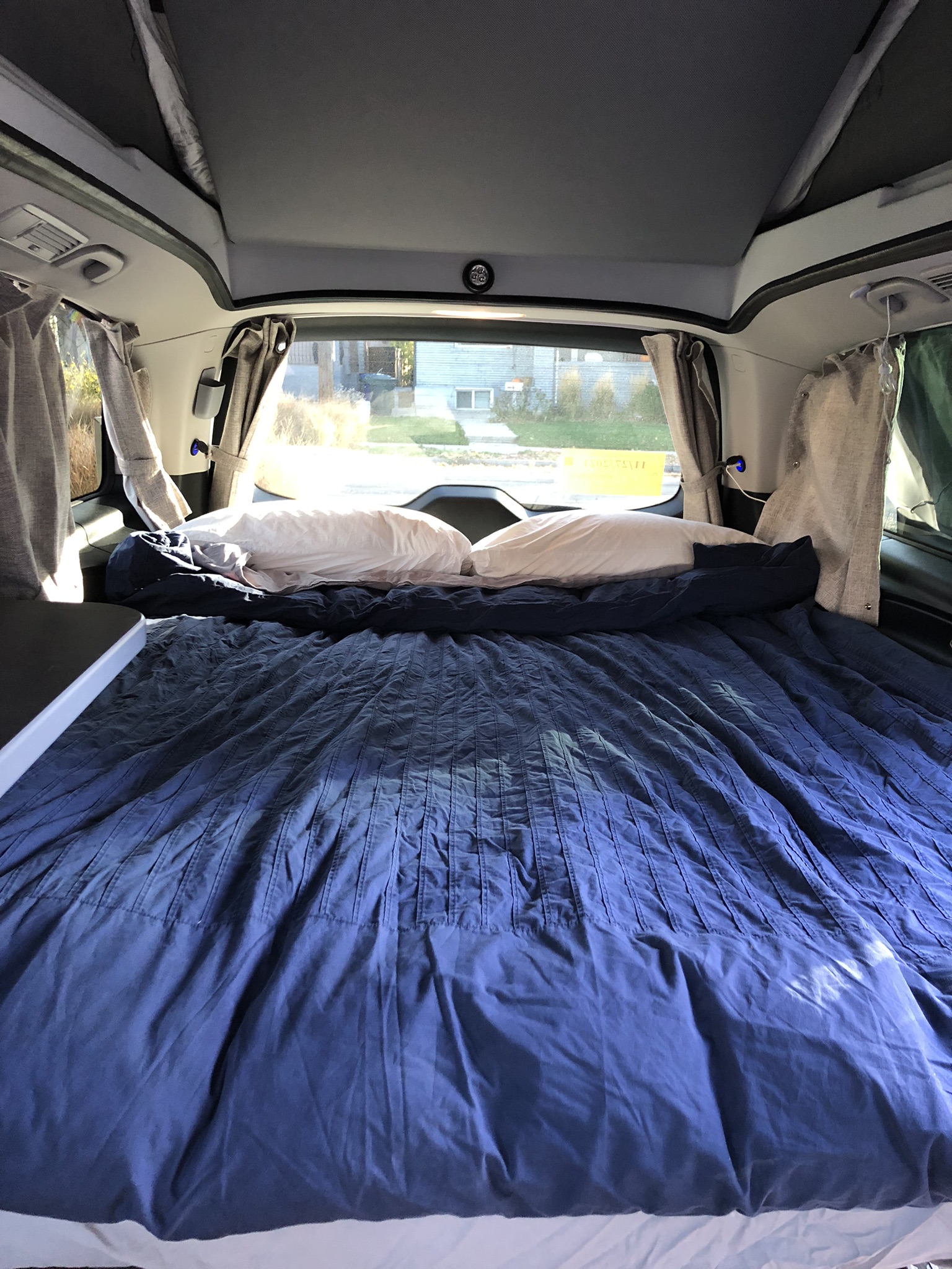 Inside a cozy RV rental: a large bed with blue bedding and pillows, curtains, and a view outside, ideal for campervan adventures.. Mercedes-Benz Metris 2021