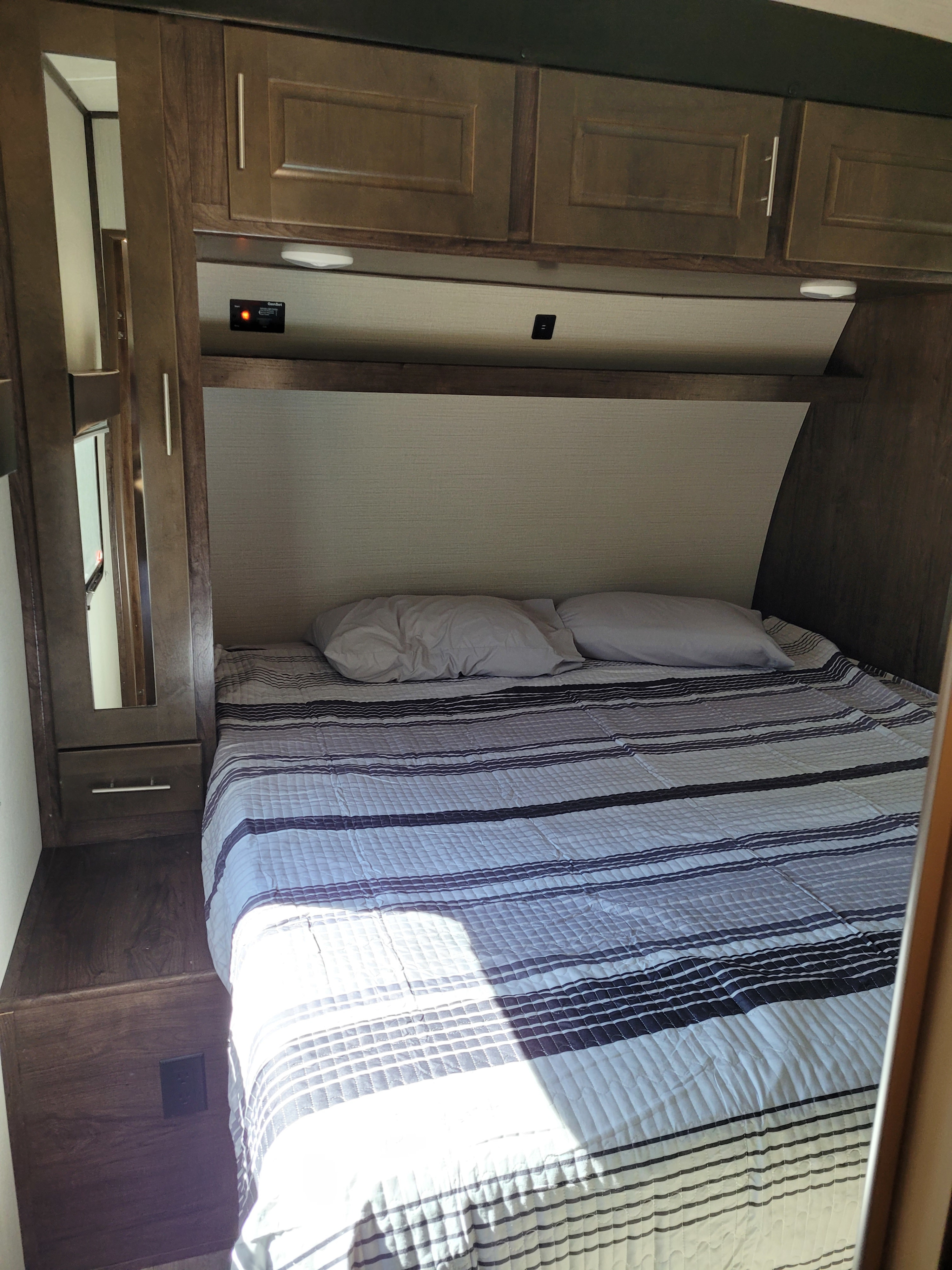 Inside an RV rental, a compact bedroom with a striped bed, pillows, overhead cabinets, and built-in storage.. Heartland Fuel 305 2018