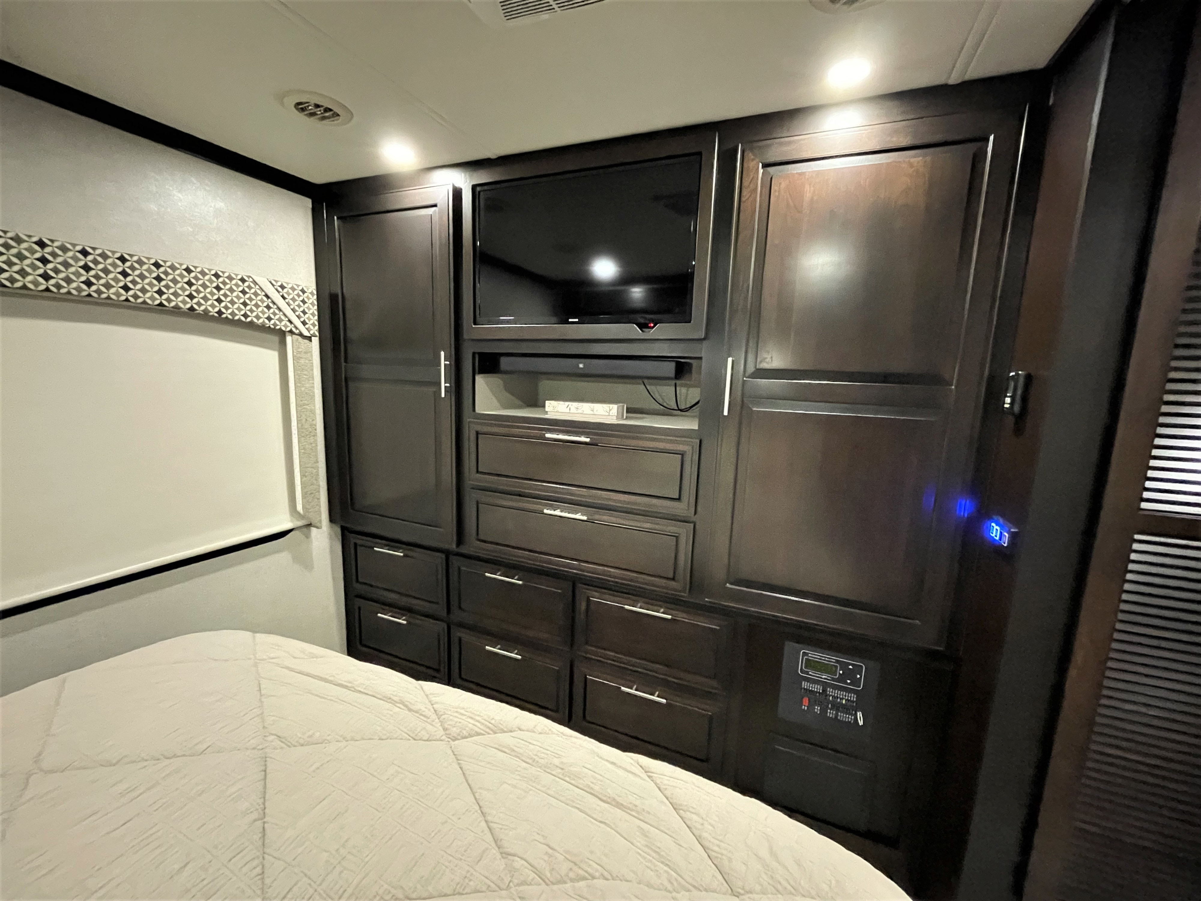 Well-appointed RV rental bedroom with a large bed, dark wood built-in unit, TV, various storage, and an RV control panel.. Dynamax Corp Dx3 2020