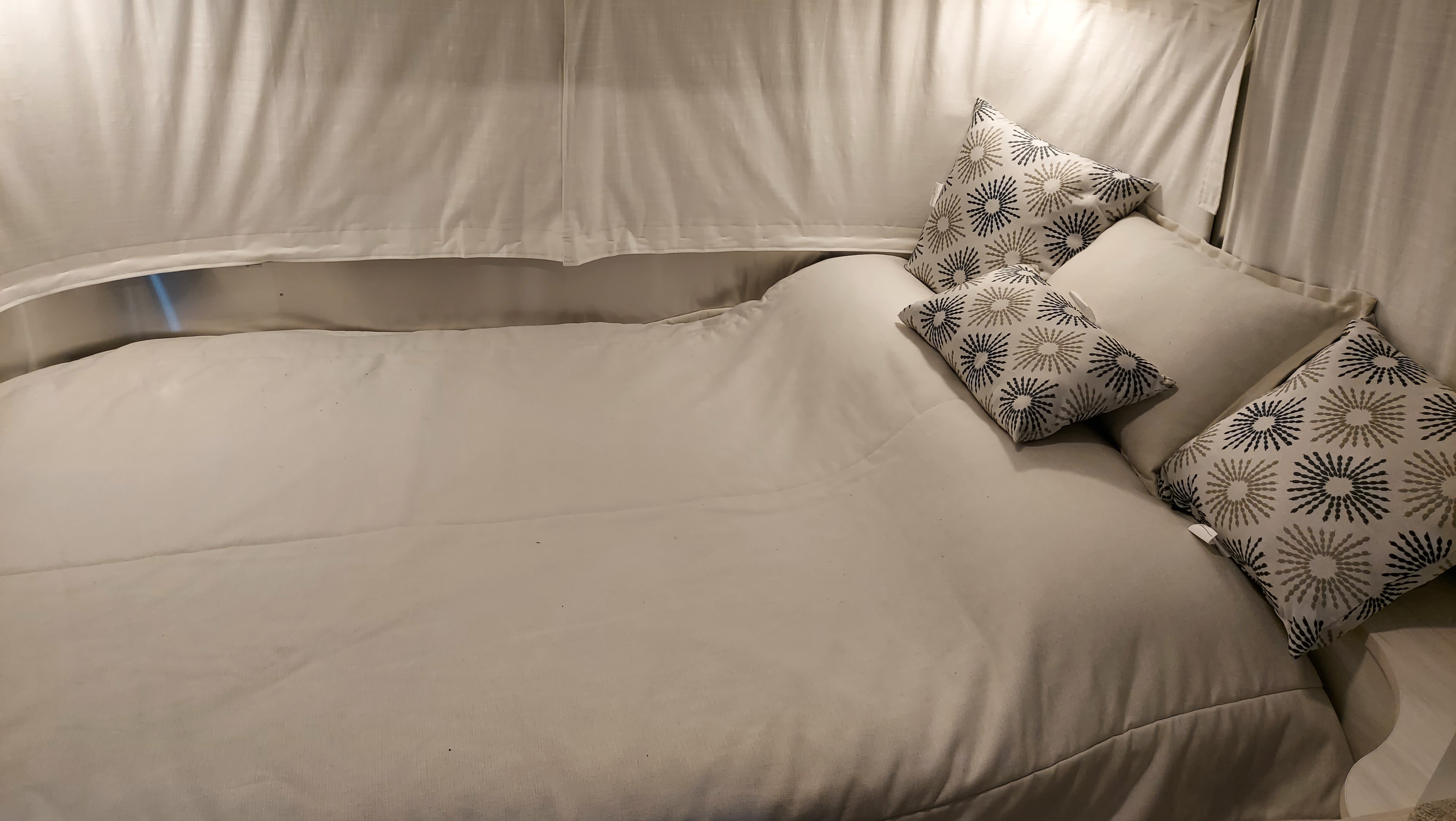 Serene RV rental sleeping area features a clean, comfortable bed with patterned pillows and light fabric walls.. Airstream Bambi 2021