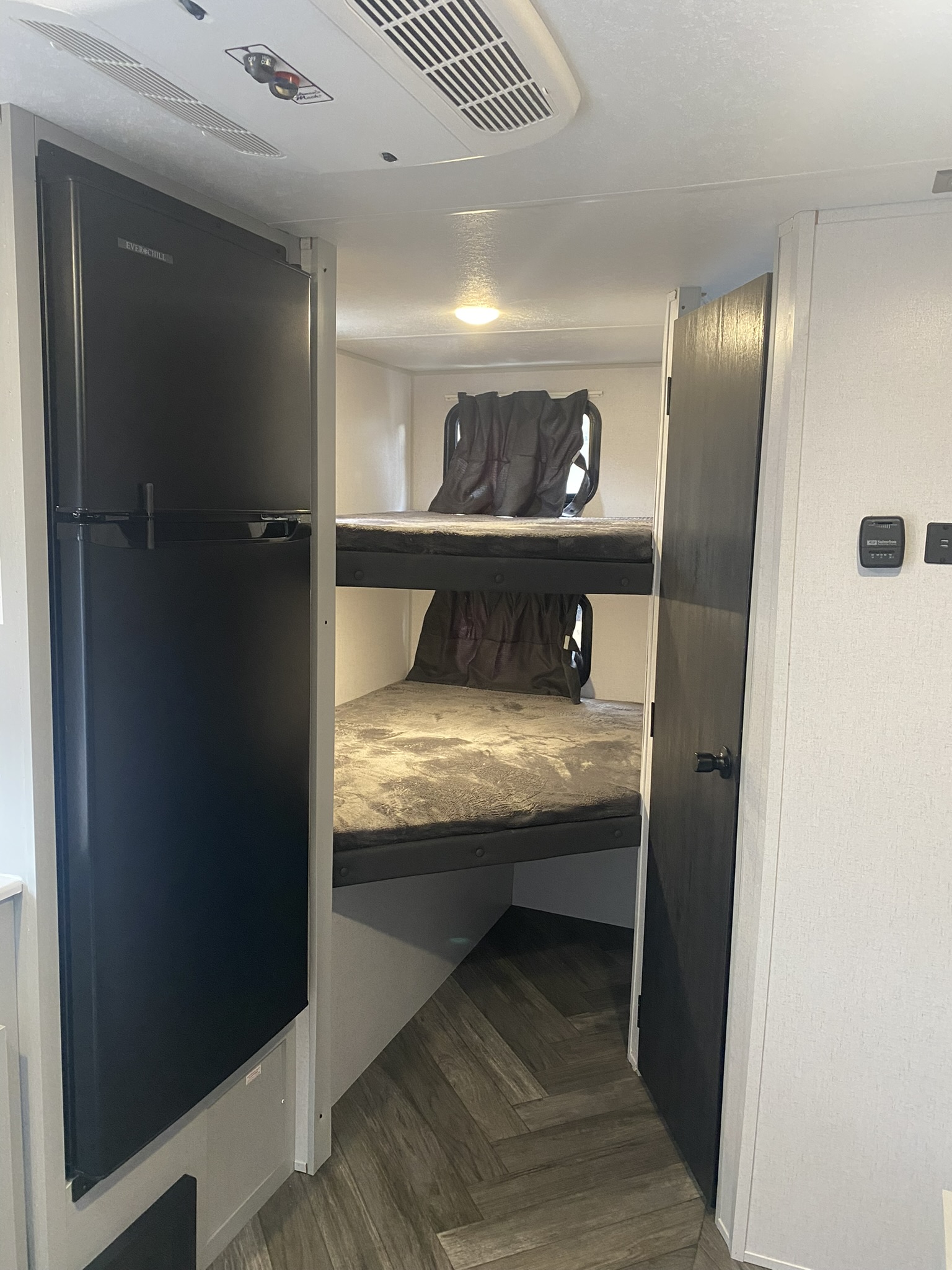 Inside an RV rental, showing a large black refrigerator on the left and bunk beds with grey bedding on the right.. Other Other 2021
