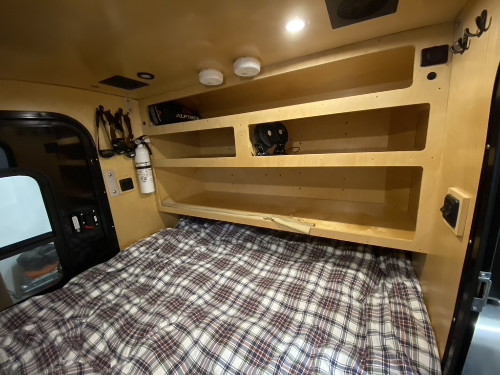 This compact RV rental features a cozy plaid bed, built-in wooden shelving, and essential safety items for travel.. Teardrop Camping Company Teardrop 2021