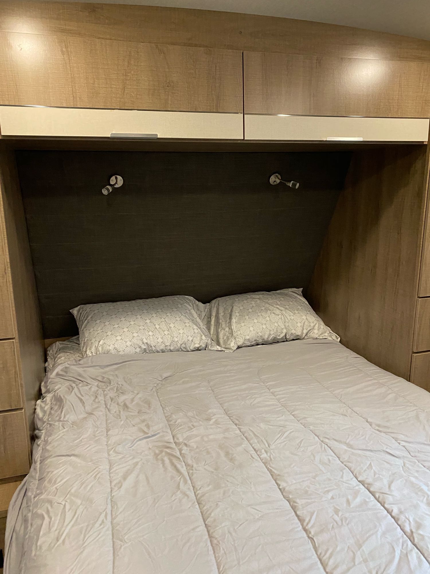 RV rental bedroom: Cozy bed with light grey bedding, pillows, reading lights, and wood-grain overhead cabinets.. Grand Design Imagine 2800BH #1 2021
