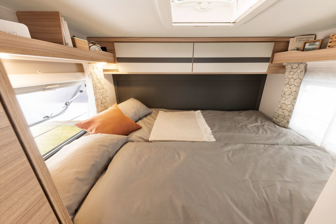 RV rental's cozy bedroom features a bed with gray linens, overhead storage, and natural light from a bright window.. Dethleffs Globebus I 1 2021