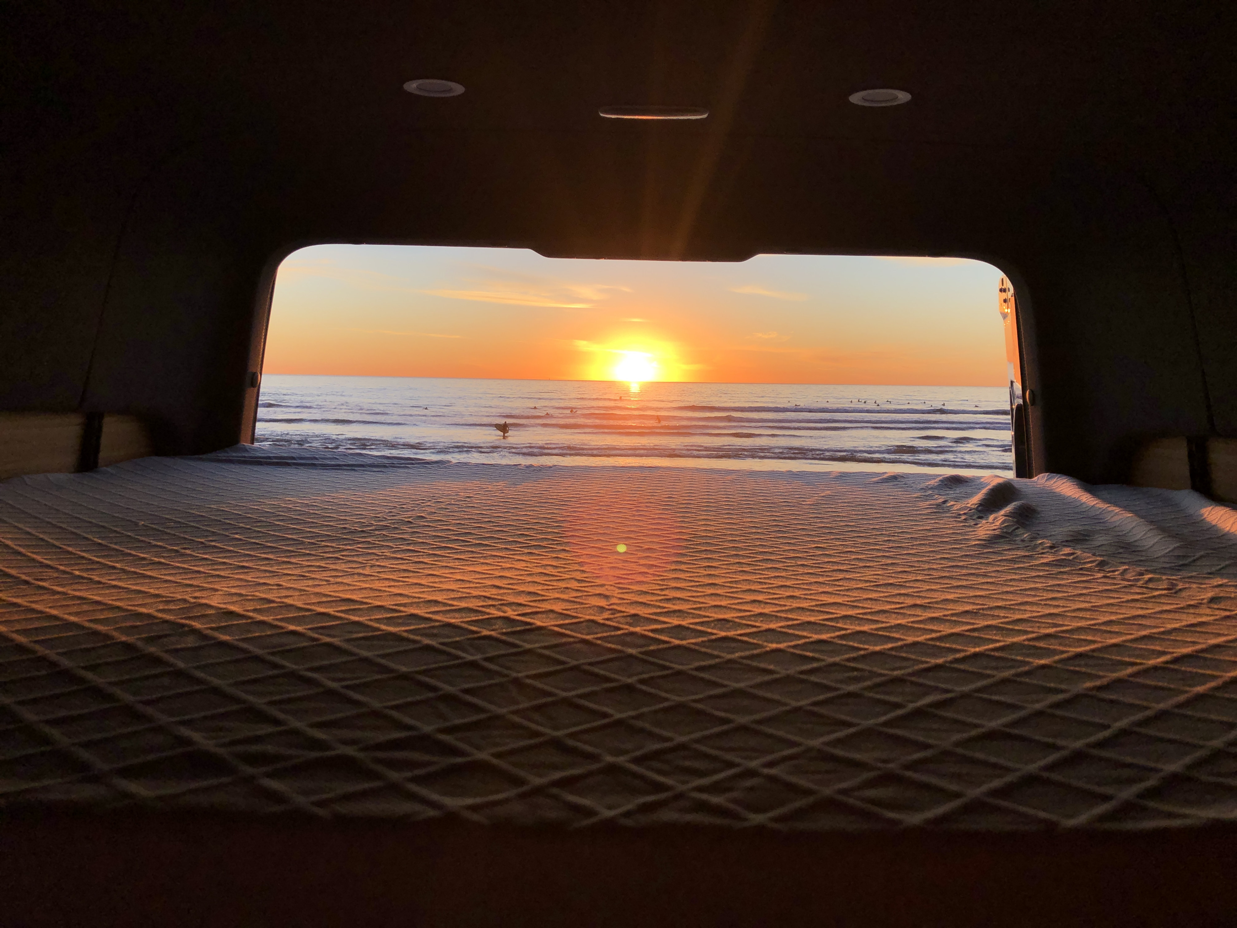 Serene RV rental view: Golden sunset over the ocean, surfers, and a comfy quilted bed inside a camper van.. Mercedes-Benz Sprinter 2021