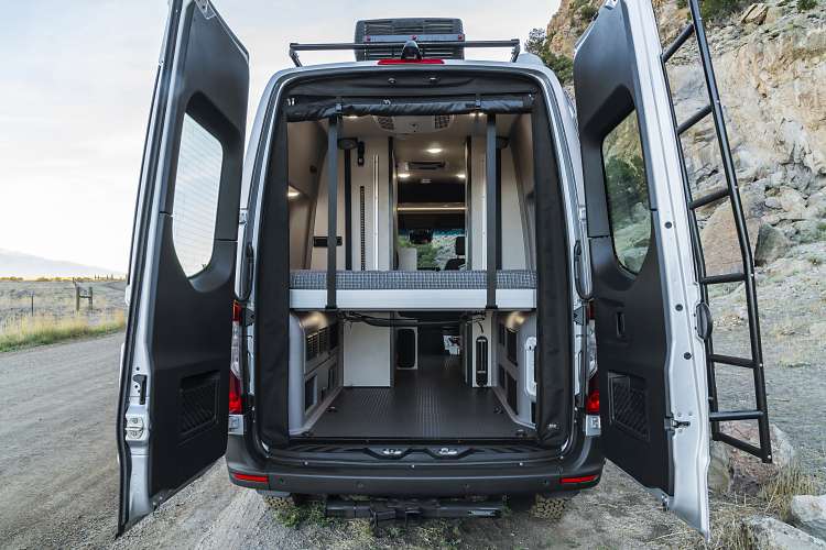 Open rear doors of an RV rental reveal a versatile interior with a raised bed over spacious gear storage, ready for adventure.. Winnebago Revel 2023