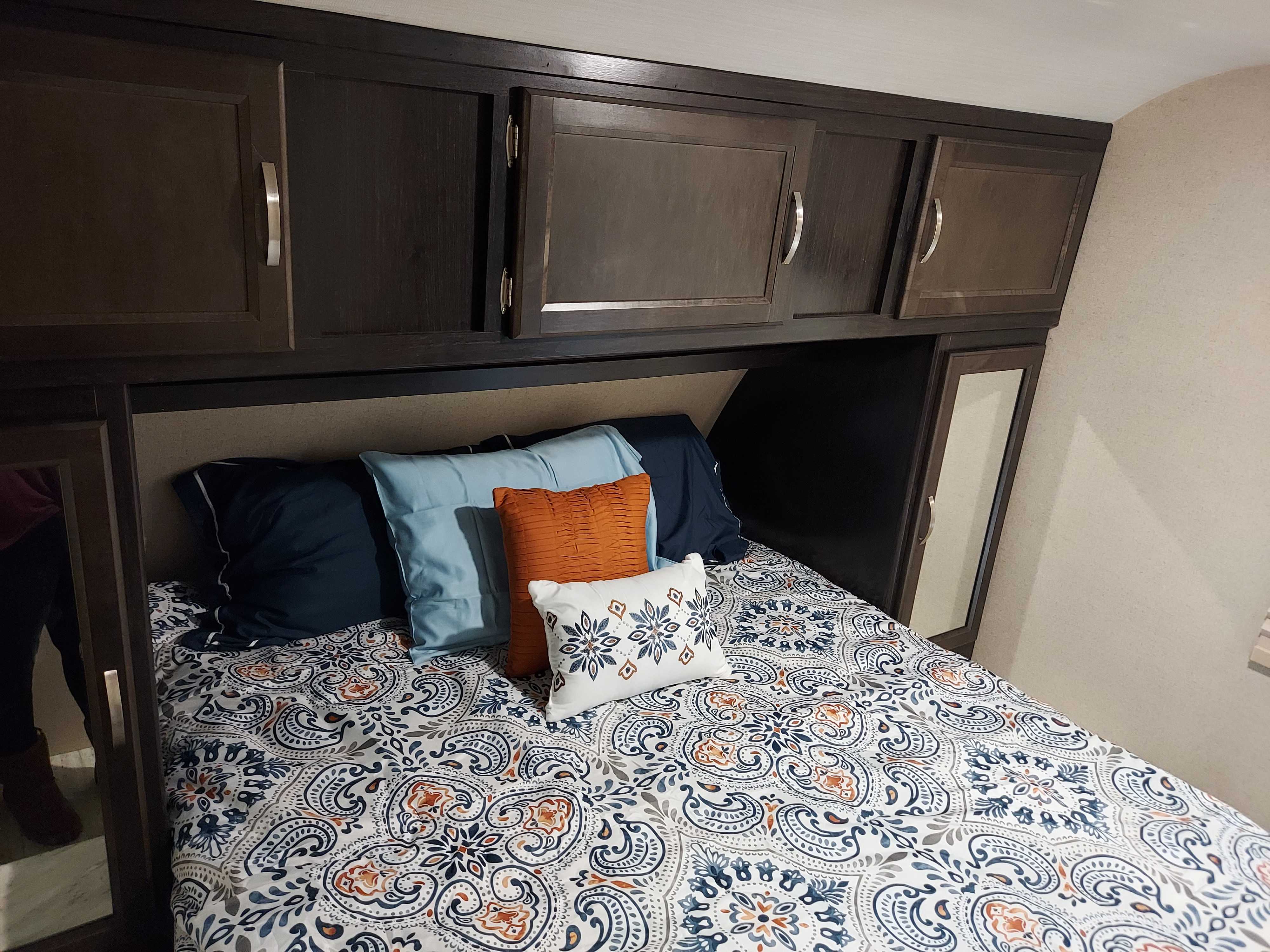 Cozy RV rental bedroom featuring a bed with a patterned comforter, decorative pillows, and dark wood overhead cabinets.. Coachmen Other 2020