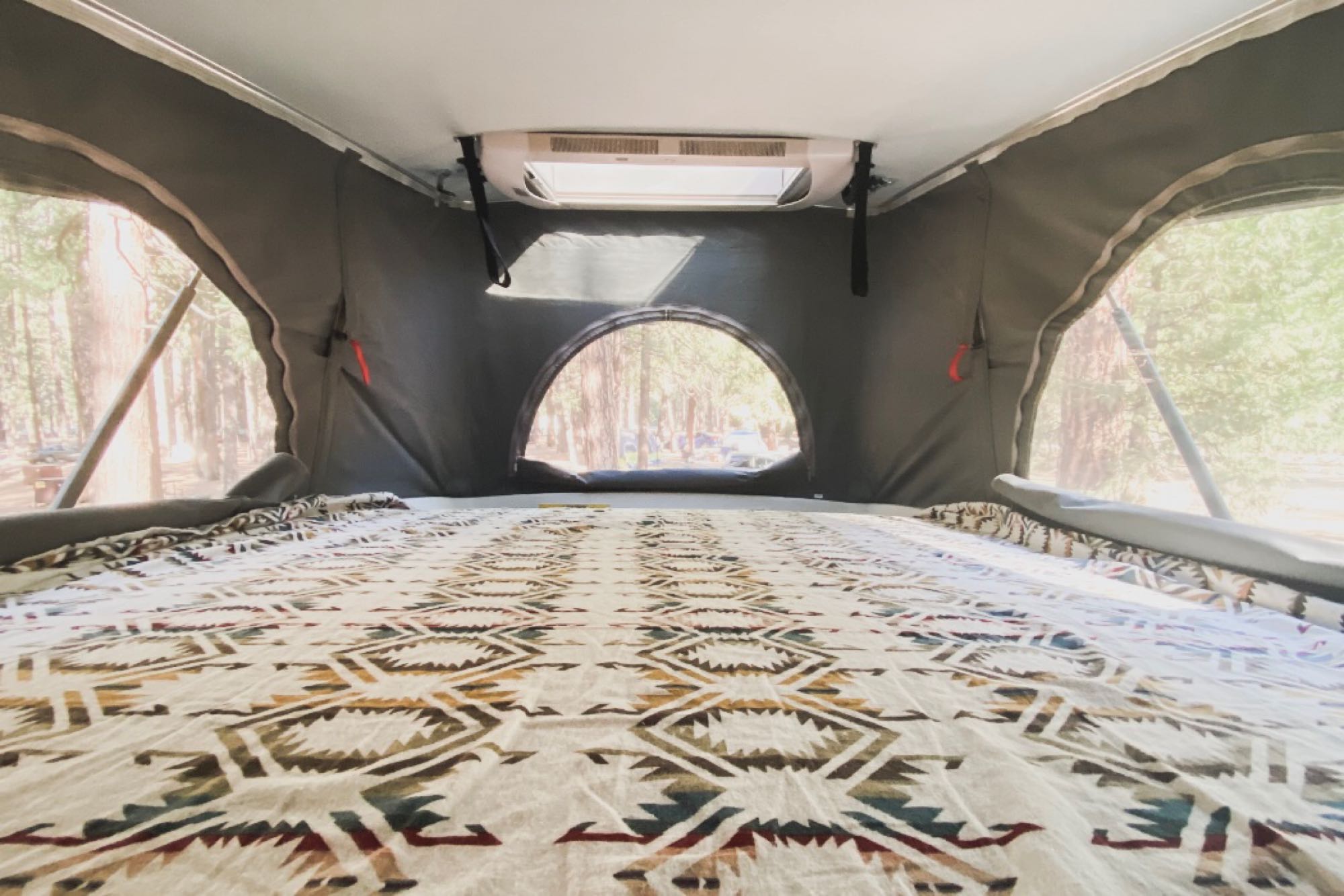 Inside an RV rental: a spacious pop-top bed with a Southwestern blanket and mesh windows overlooking a forest campsite.. Hymer Hymer Class B 2019
