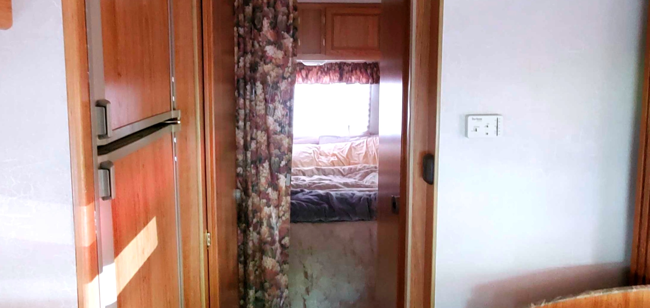 RV rental interior: A wooden refrigerator, floral curtain, and a view into a compact bedroom with a window.. Coachmen Leprechaun 2000