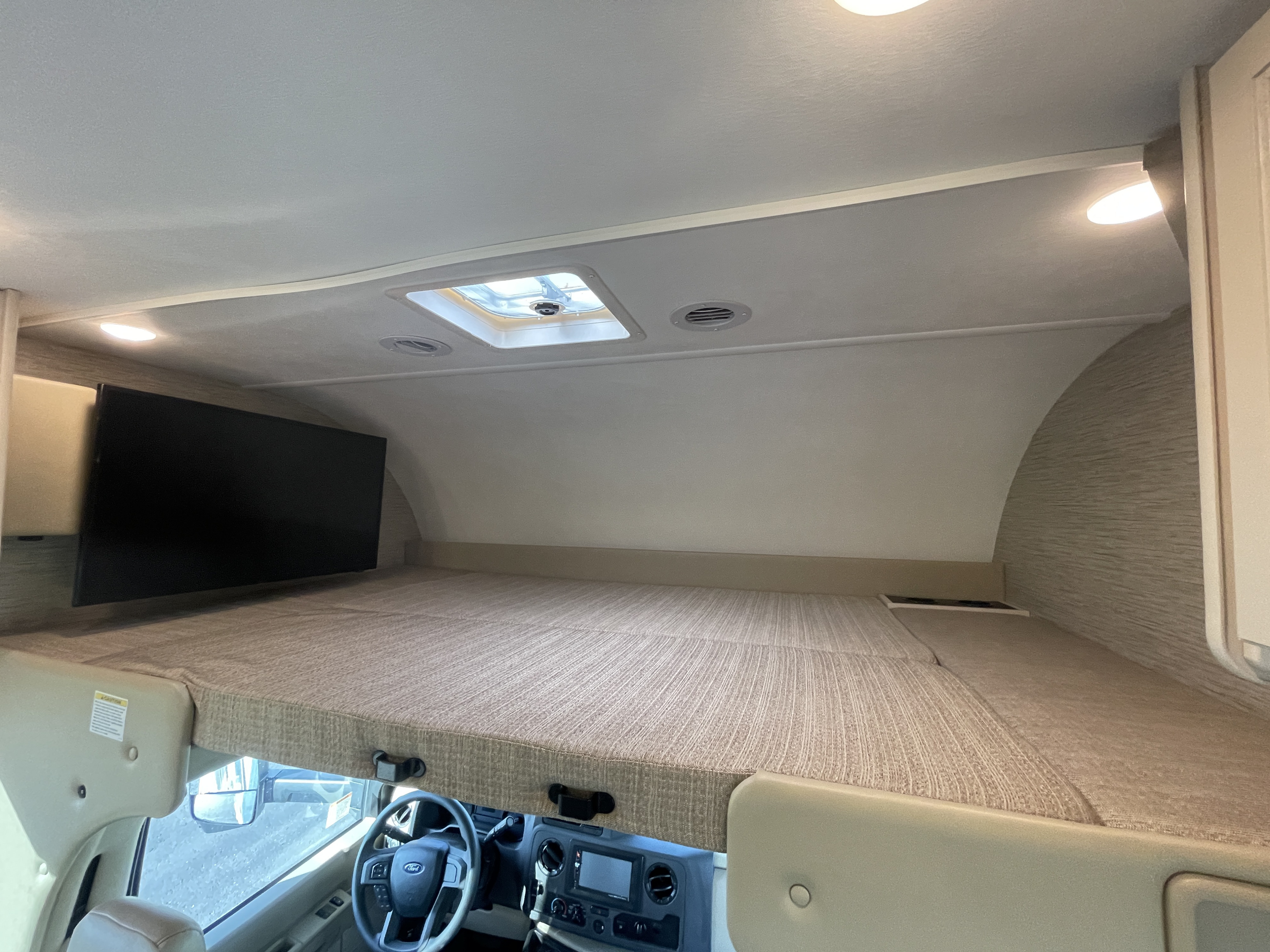 Comfortable RV rental cab-over bunk area with a large bed, mounted TV, skylight, and vehicle dashboard visible below.. Thor Motor Coach Four Winds 2022
