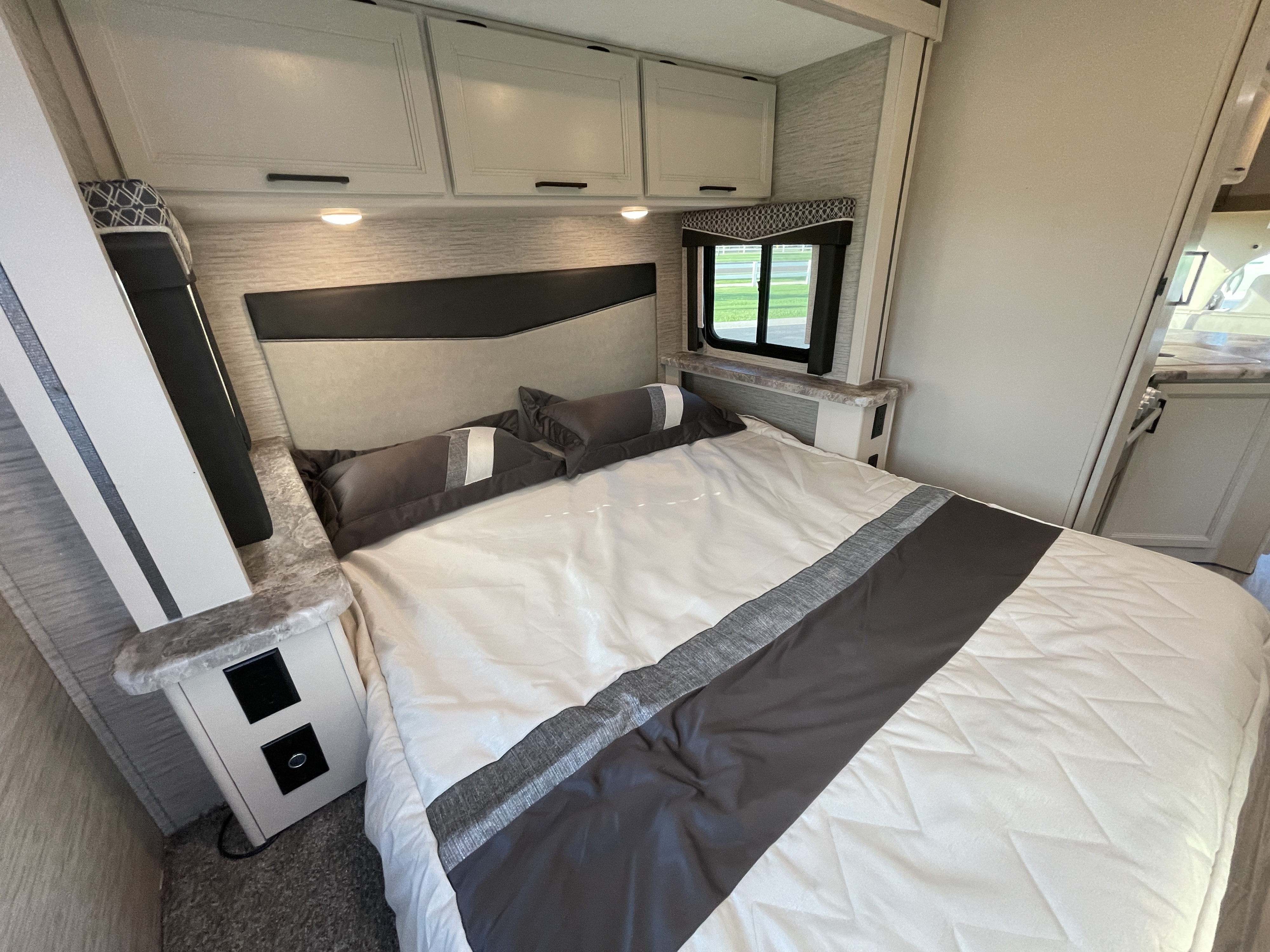 Spacious RV rental bedroom featuring a full bed with gray and white bedding, overhead cabinets, and a window.. Thor Motor Coach Four Winds 2022