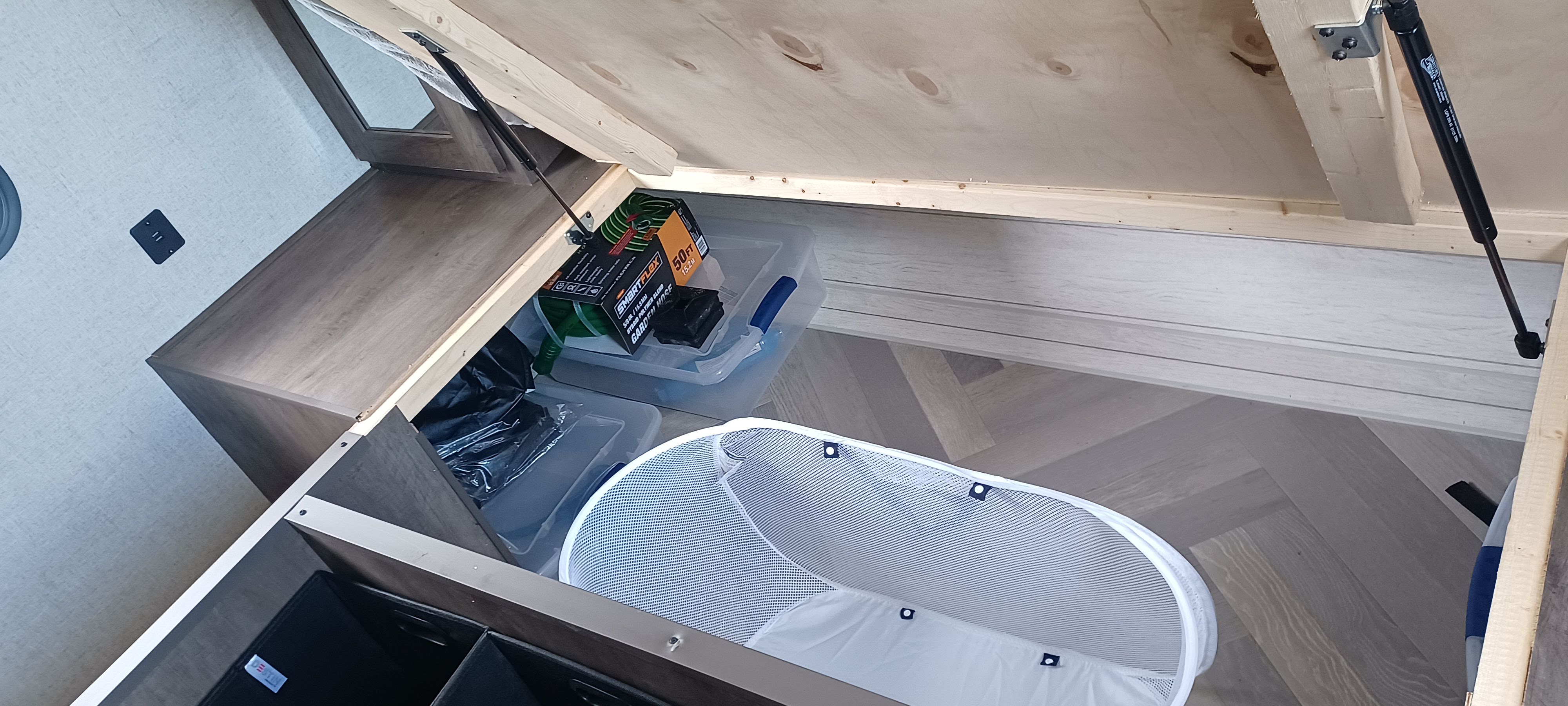RV rental bed lifted for under-bed storage: clear bins, garden hose, and a mesh basket on light wood flooring.. Forest River Other 2021