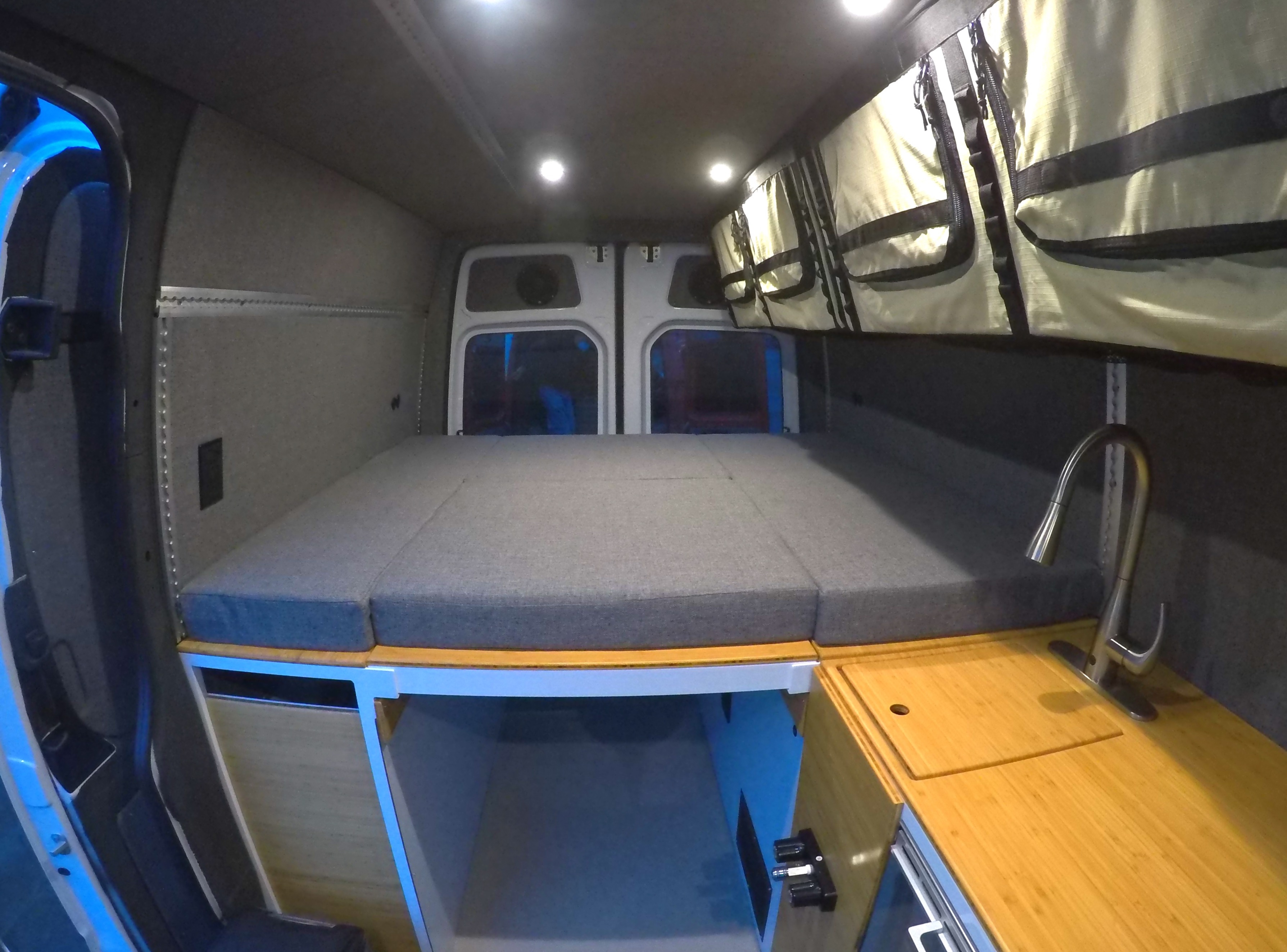 Spacious rv rental camper van interior featuring a comfortable gray bed, compact kitchen with a sink, and overhead storage bags.. Mercedes Benz Sprinter 2018
