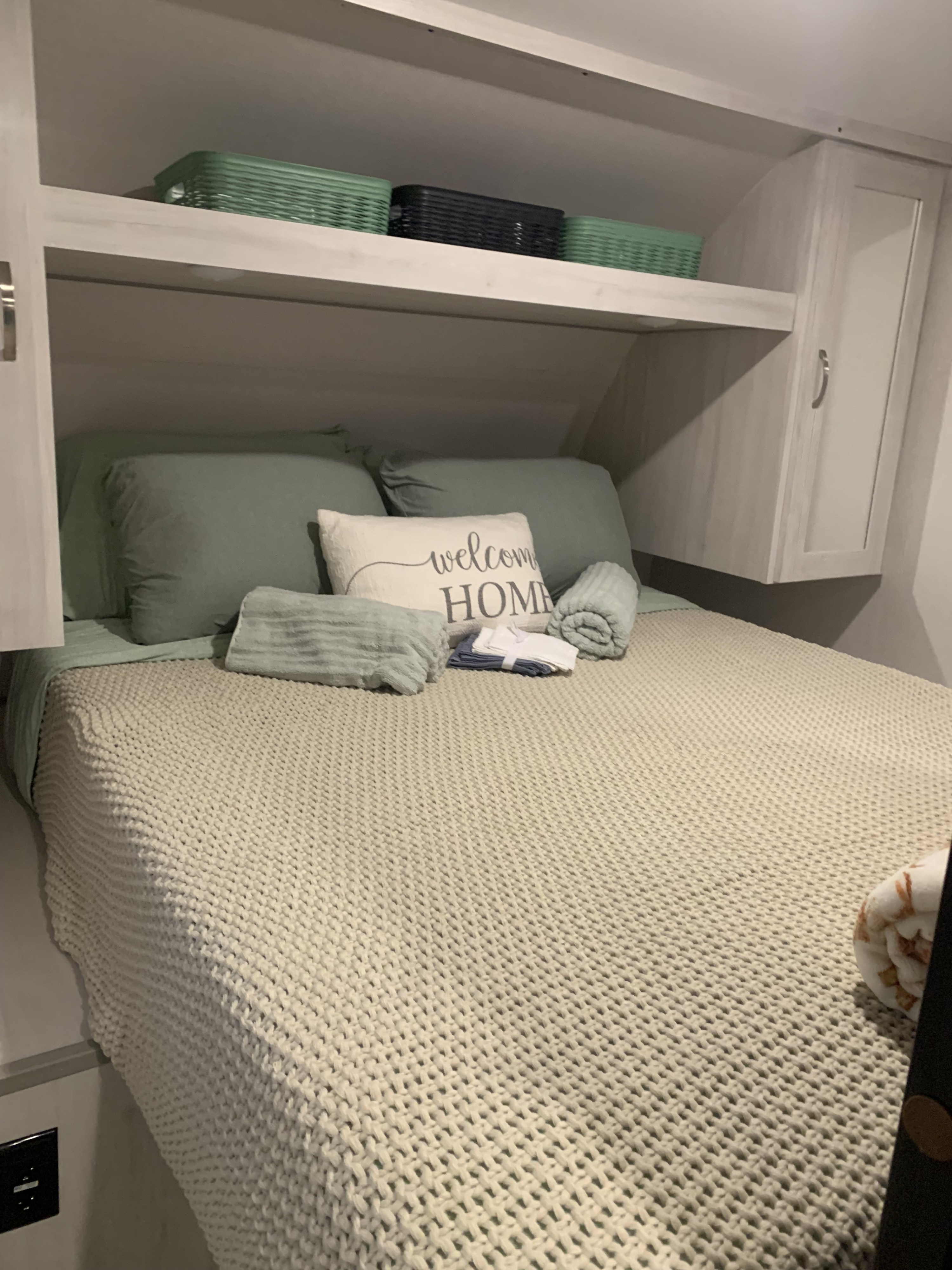 Comfy bed in an RV rental with "Welcome HOME" pillow, neatly folded towels, and storage baskets on shelves, creating a welcoming bedroom.. Prime Time Tracer 2022
