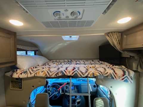 Comfortable cab-over bunk in an RV rental, featuring a bed, overhead AC, and view into the driver's cockpit.. Thor Freedom Elite 2021