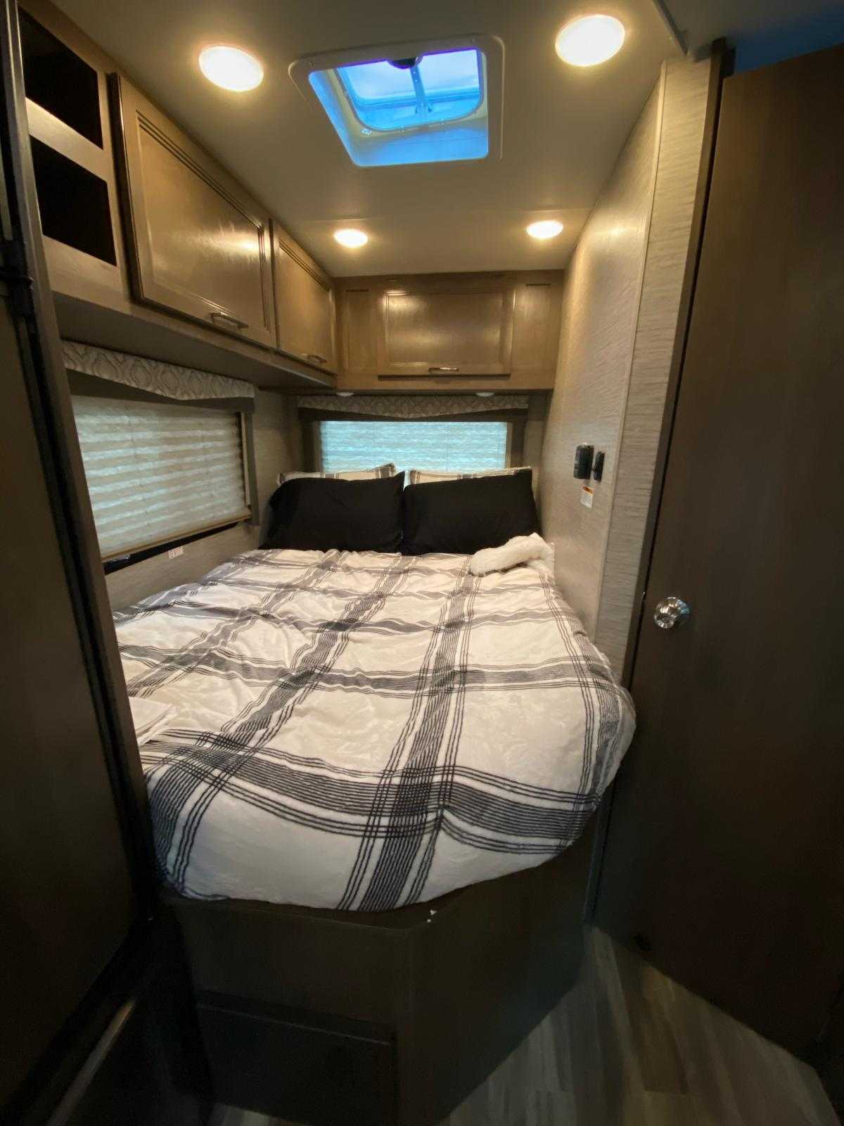 A cozy RV rental bedroom features a plaid bed, overhead wood-tone cabinets, and bright ceiling lights with a skylight.. Thor Freedom Elite 2021