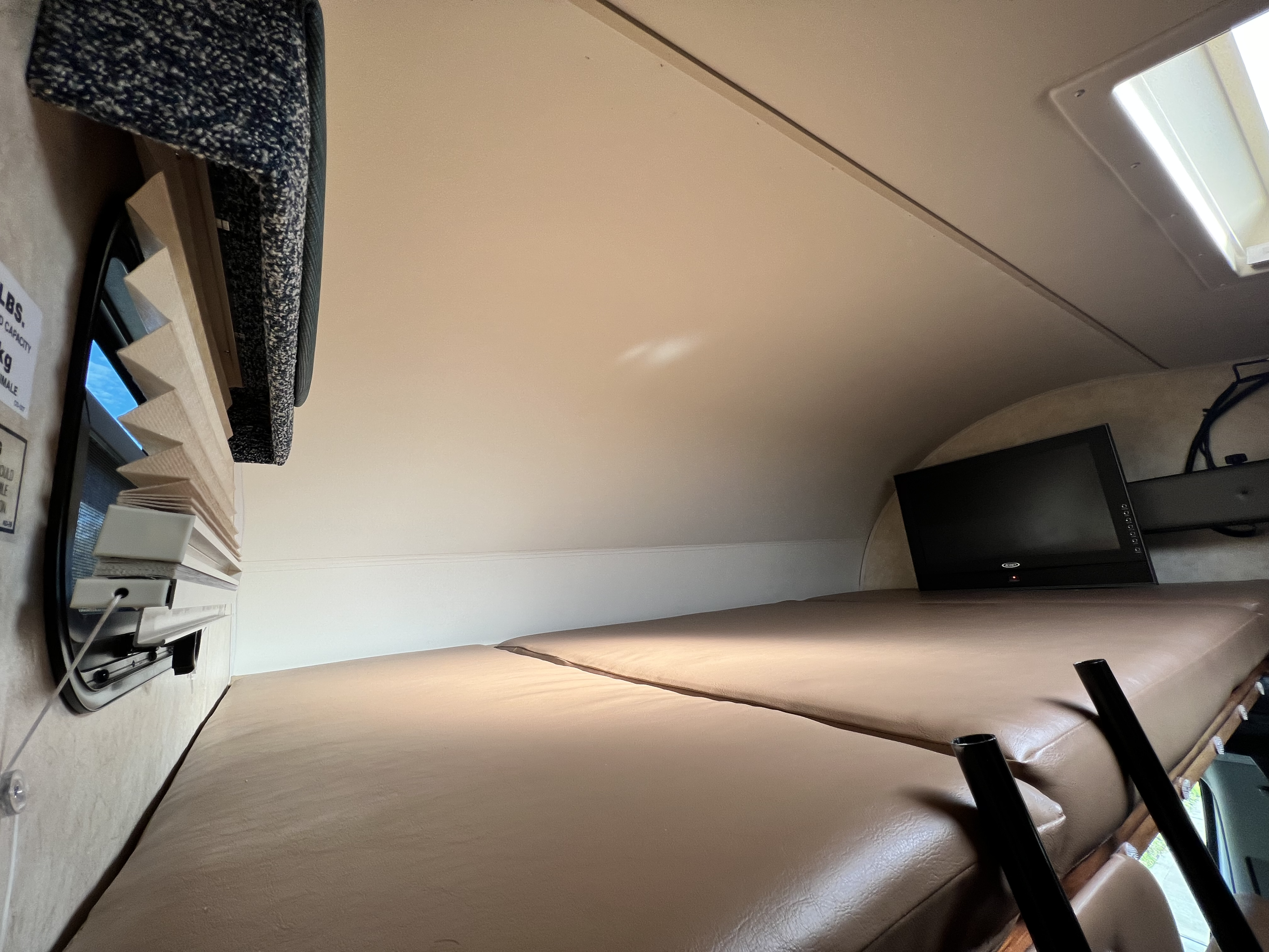 Inside an RV rental: a cozy overhead bunk bed with mattress, TV, window shades, and a skylight above.. Winnebago Mercedes 2012