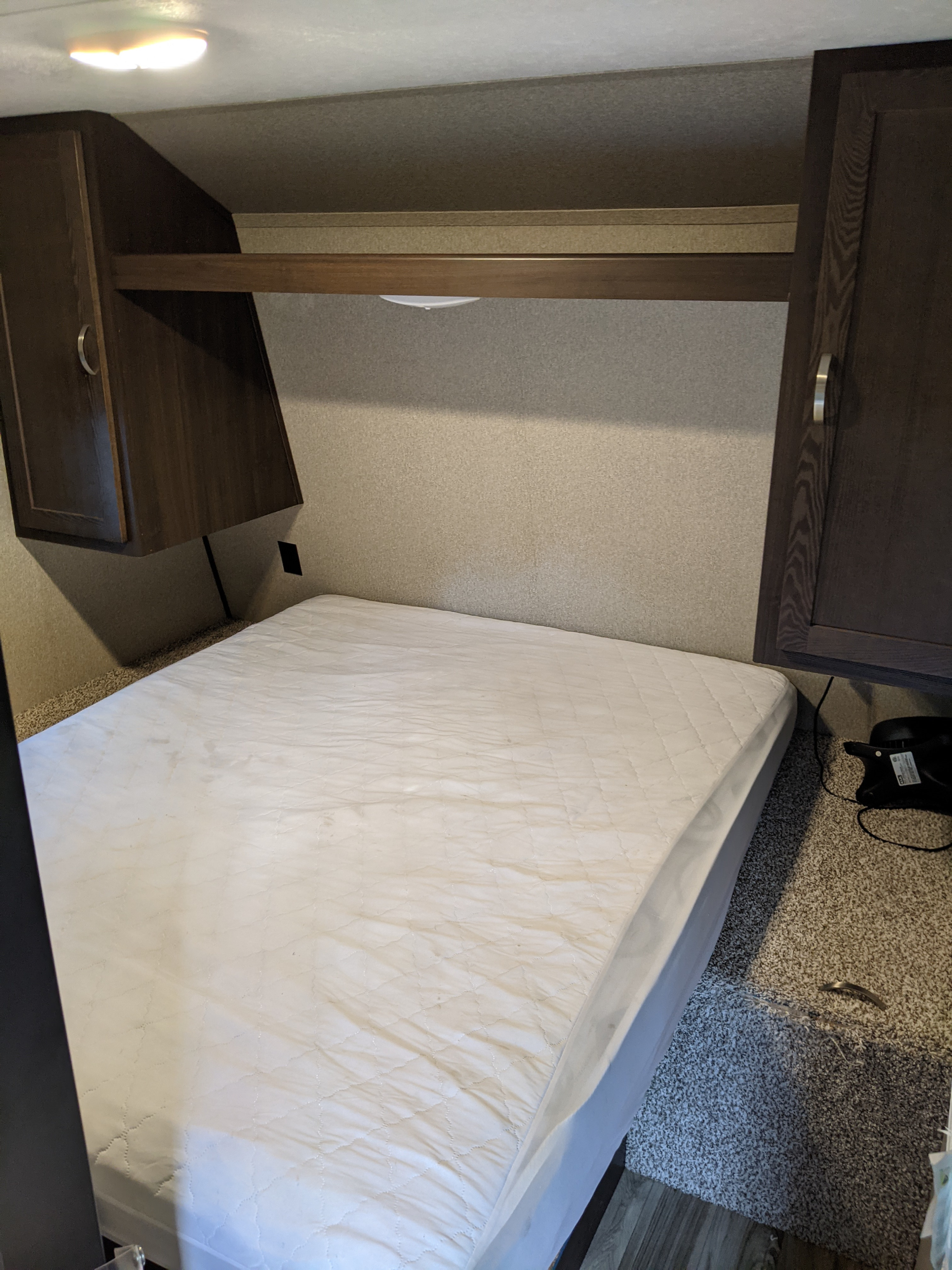 Inside an RV rental, a compact bedroom features a white mattress, overhead cabinets, a shelf, and carpeted flooring.. Keystone Springdale 2018