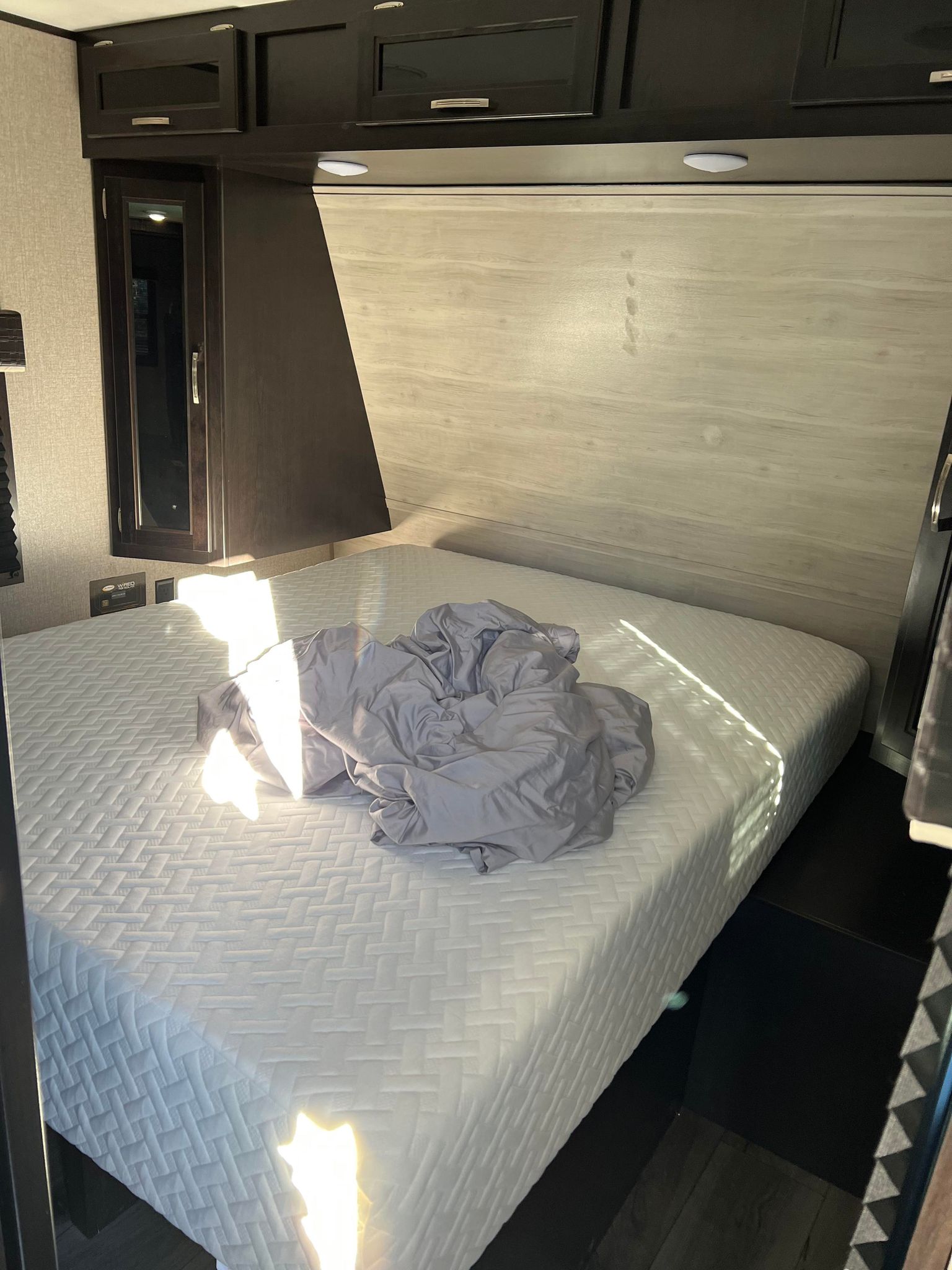 RV rental interior: cozy bedroom with a white mattress and gray bedding, illuminated by sunlight. Overhead storage and outlets visible.. Jayco Jay Flight 2021