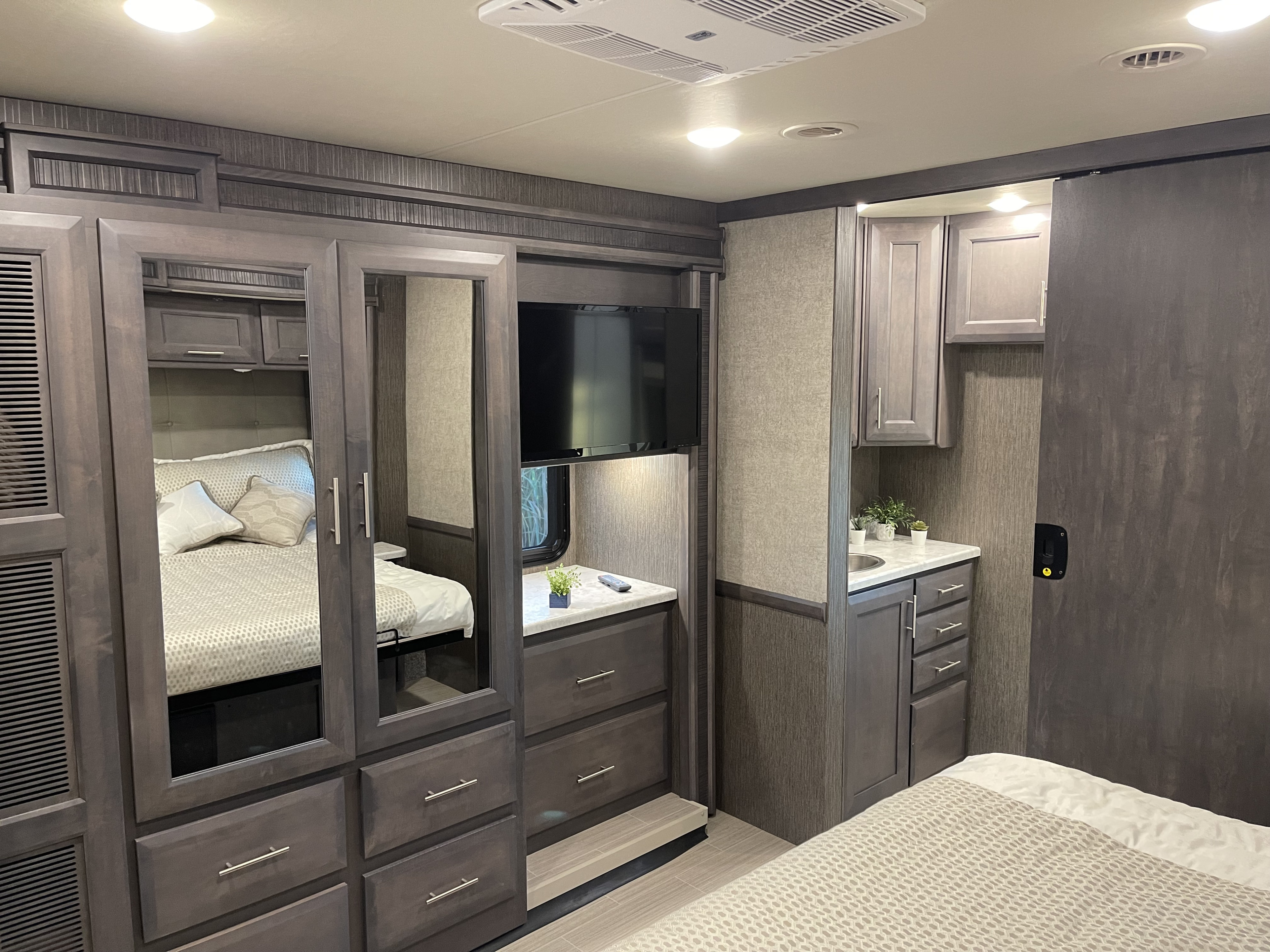 Modern RV rental bedroom featuring mirrored wardrobe, flat-screen TV, comfy bed, and compact vanity with sink.. Thor Challenger 2021