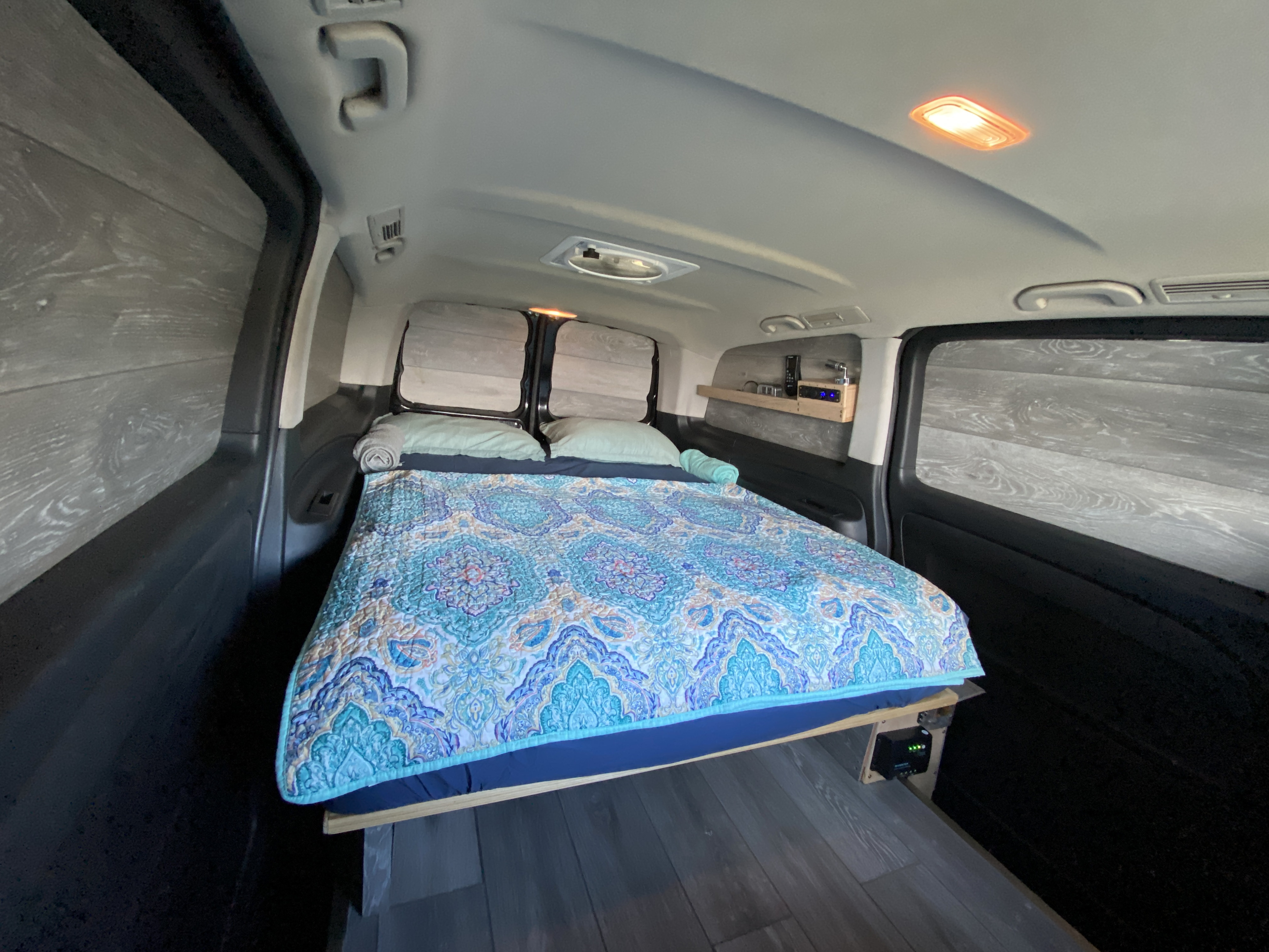 Cozy RV rental camper van interior. Features a spacious bed with a vibrant blue-patterned quilt, wood-look walls & floor.. Mercedes-Benz METRIS 2018