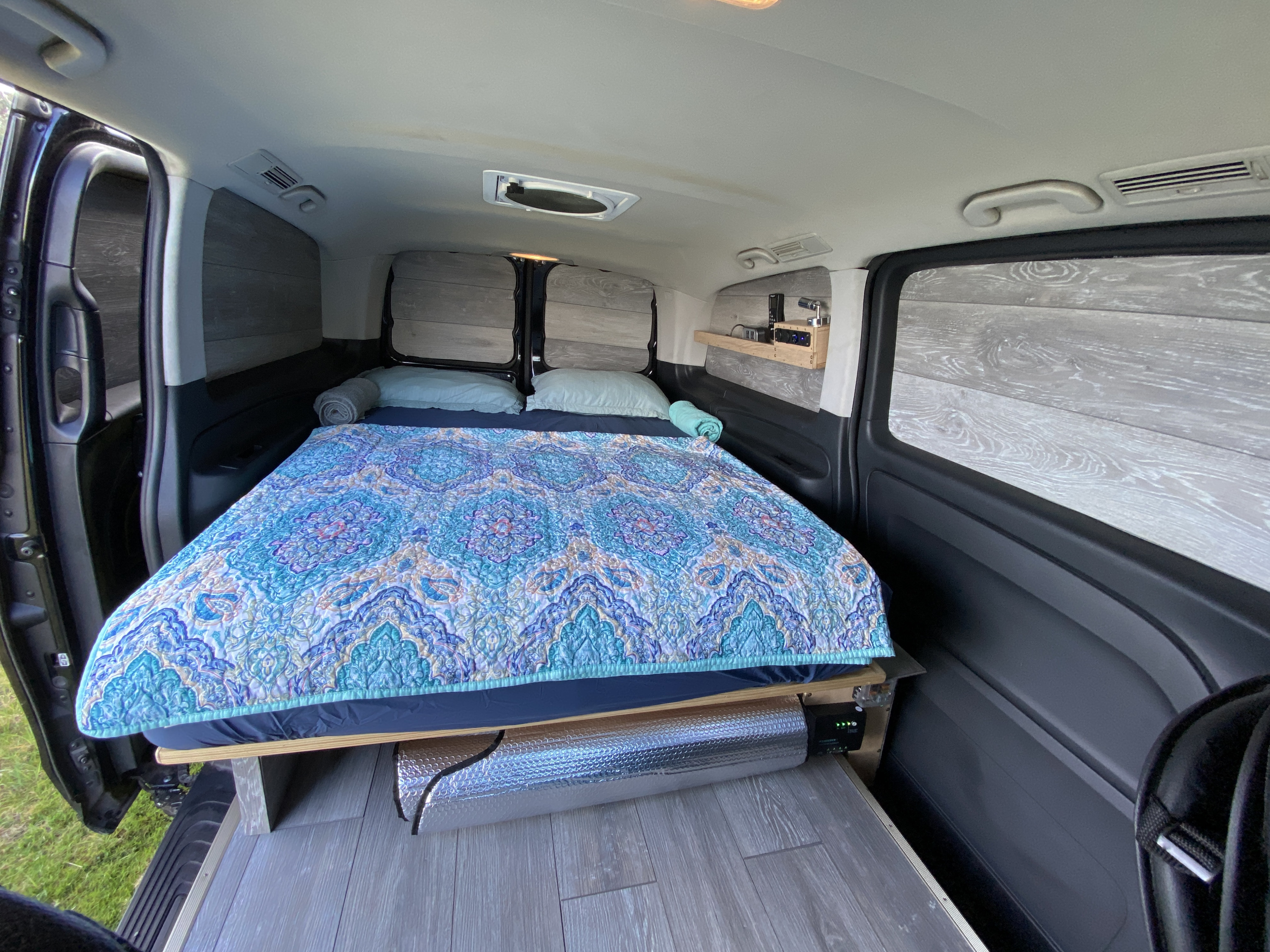 Cozy camper van interior, ideal for your next **rv rental** adventure. Features a comfy bed, wood paneling, and electronics shelf.. Mercedes-Benz METRIS 2018