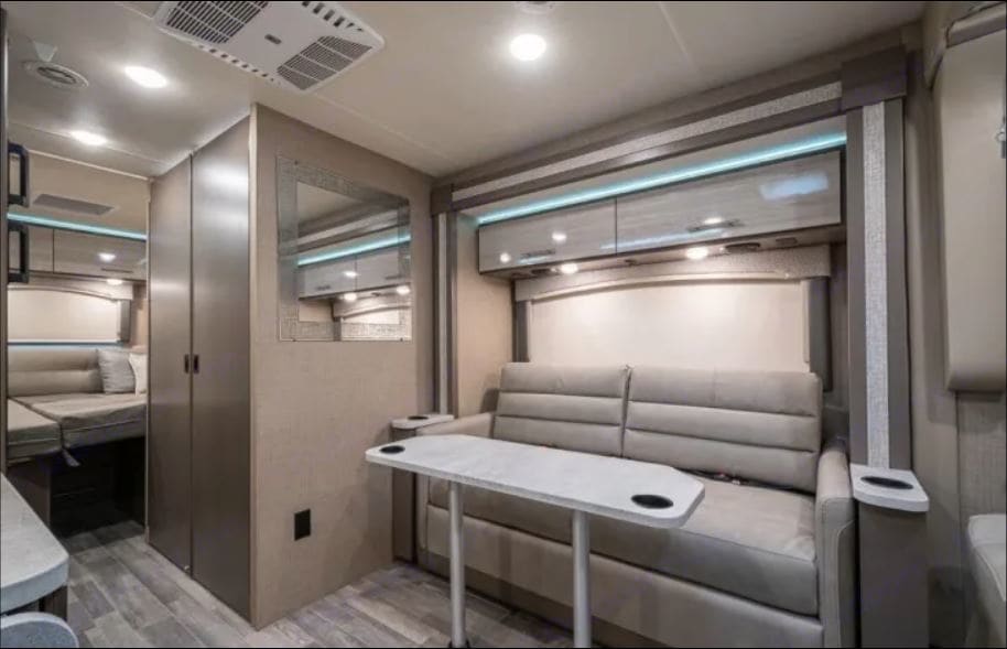 An rv rental's cozy & modern interior: living area with sofa, removable table, overhead cabinets, and a rear bed.. Thor Motor Coach Vegas 2020