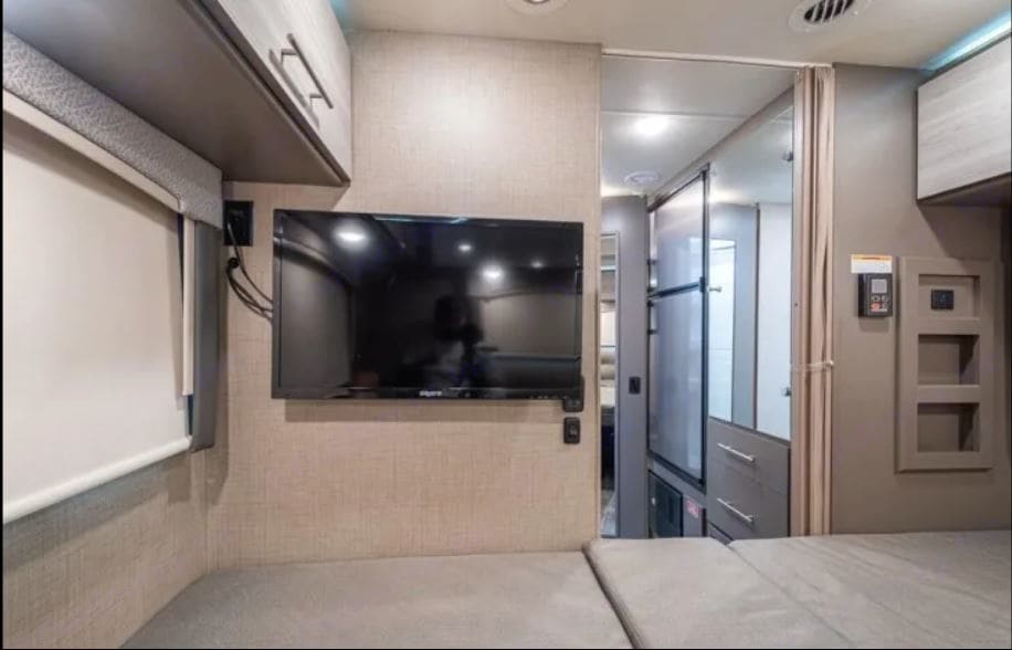 Modern RV rental interior featuring a wall-mounted TV, comfortable sleeping/lounge area, overhead storage, and control panel.. Thor Motor Coach Vegas 2020