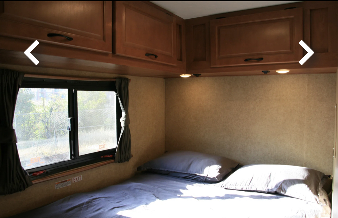 Cozy rv rental bedroom featuring a made bed, large window, and wooden overhead storage cabinets.. Thor Majestic 2017
