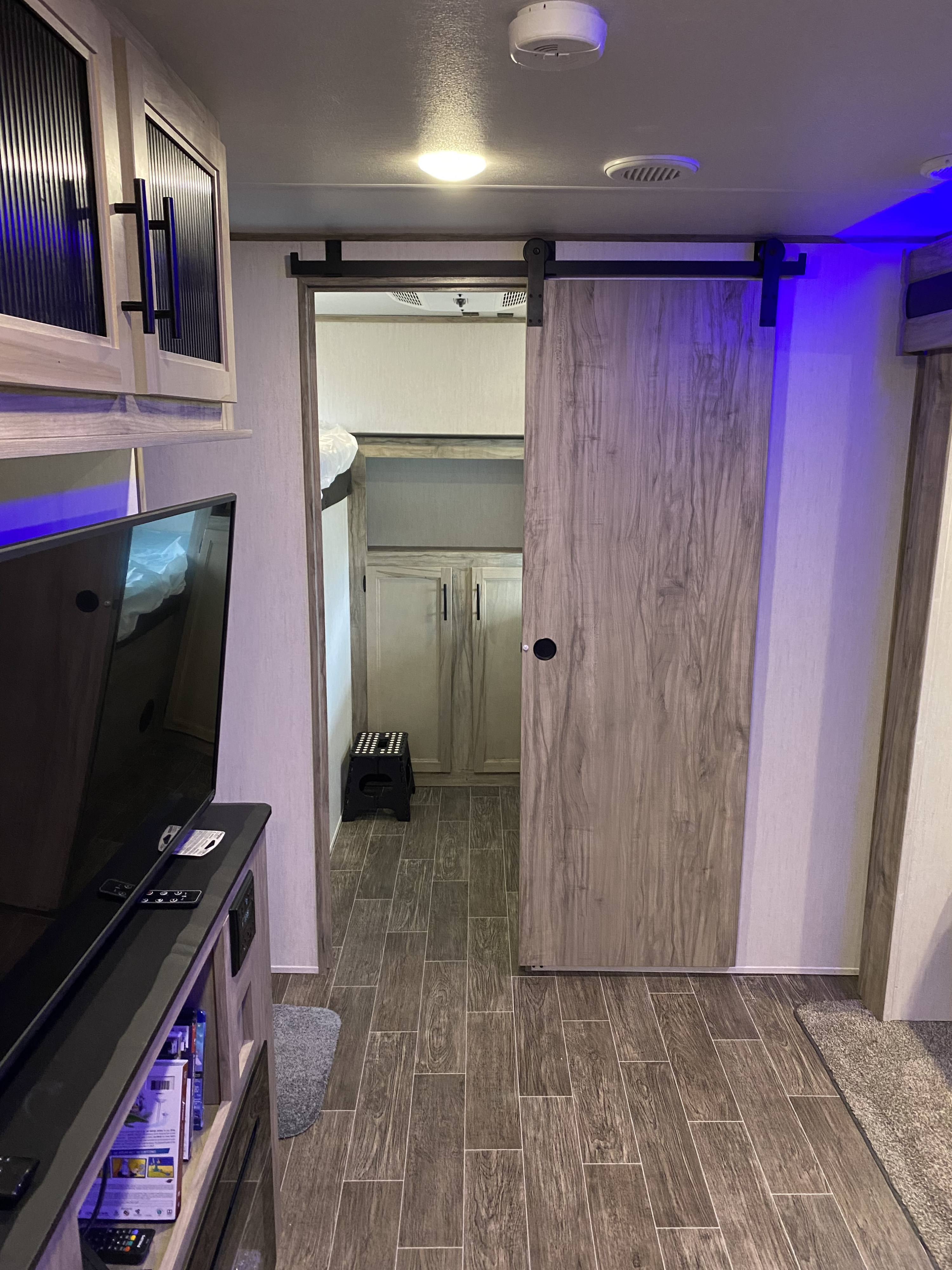 RV rental interior: Modern living area with TV, overhead storage, and a partially open barn door to a bunk room with cabinets.. Forest River Cherokee Alpha Wolf 2021