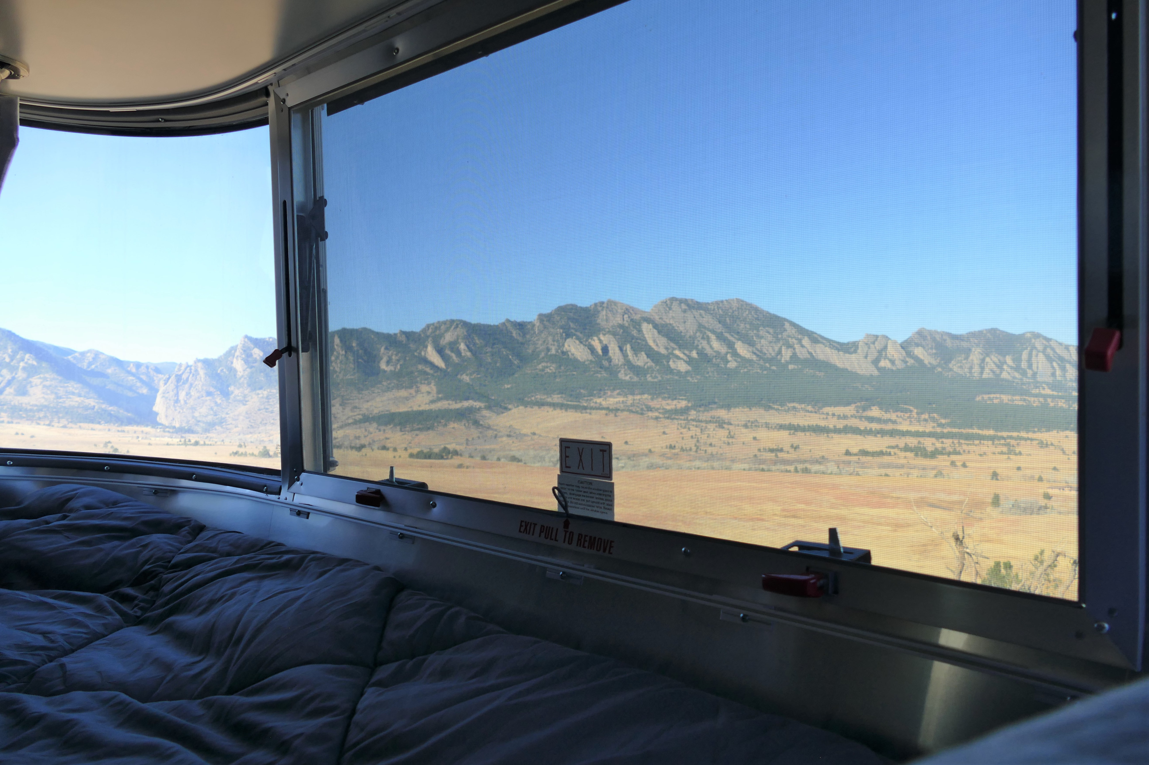 An RV rental's cozy bed and window with a screen showcasing rugged mountains and a golden prairie under a clear sky.. Airstream Caravel 2020