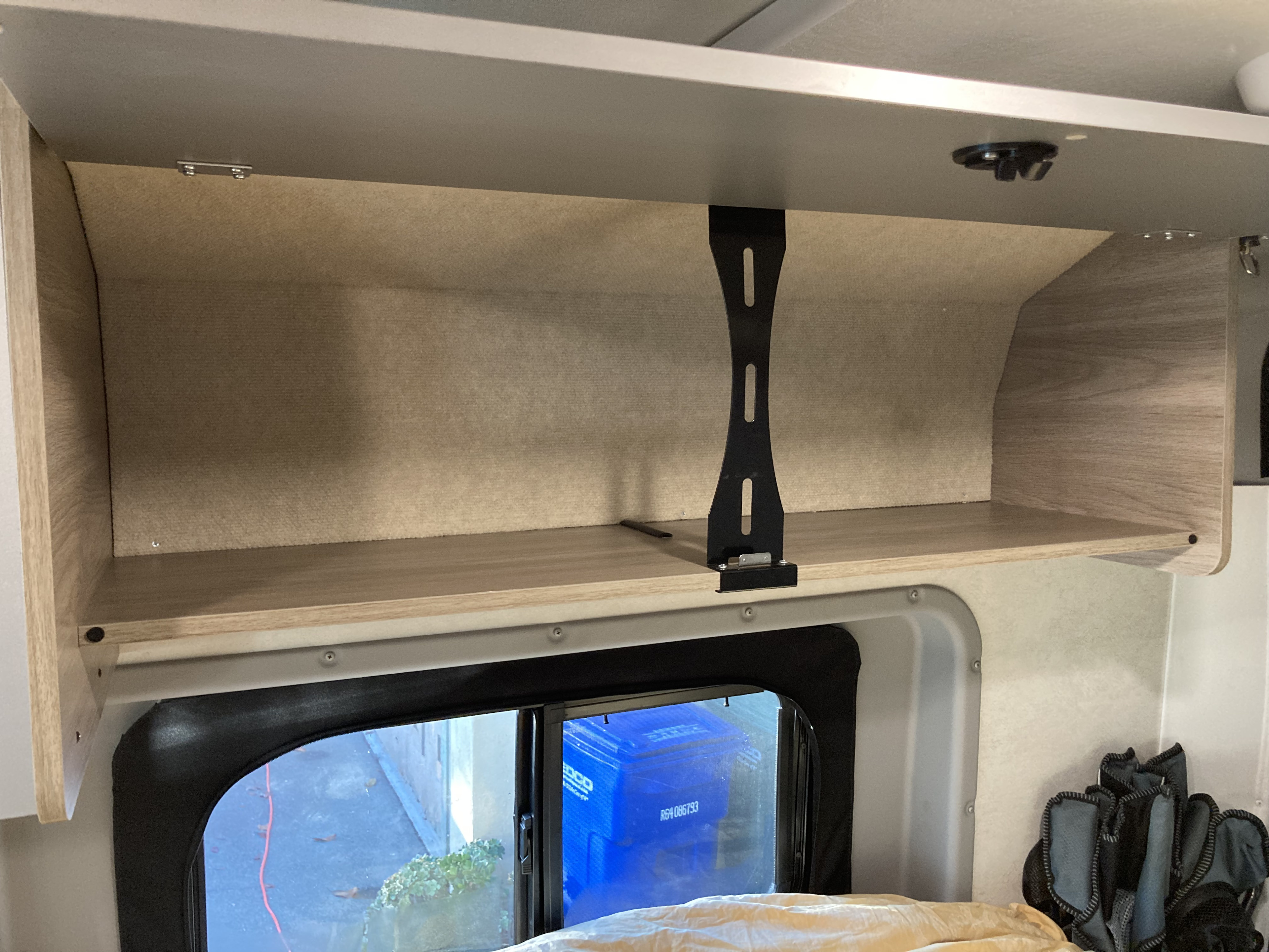 An rv rental's overhead cabinet and shelf, seen from inside, with a window below showing outdoors.. Winnebago Solis 59PX 2022
