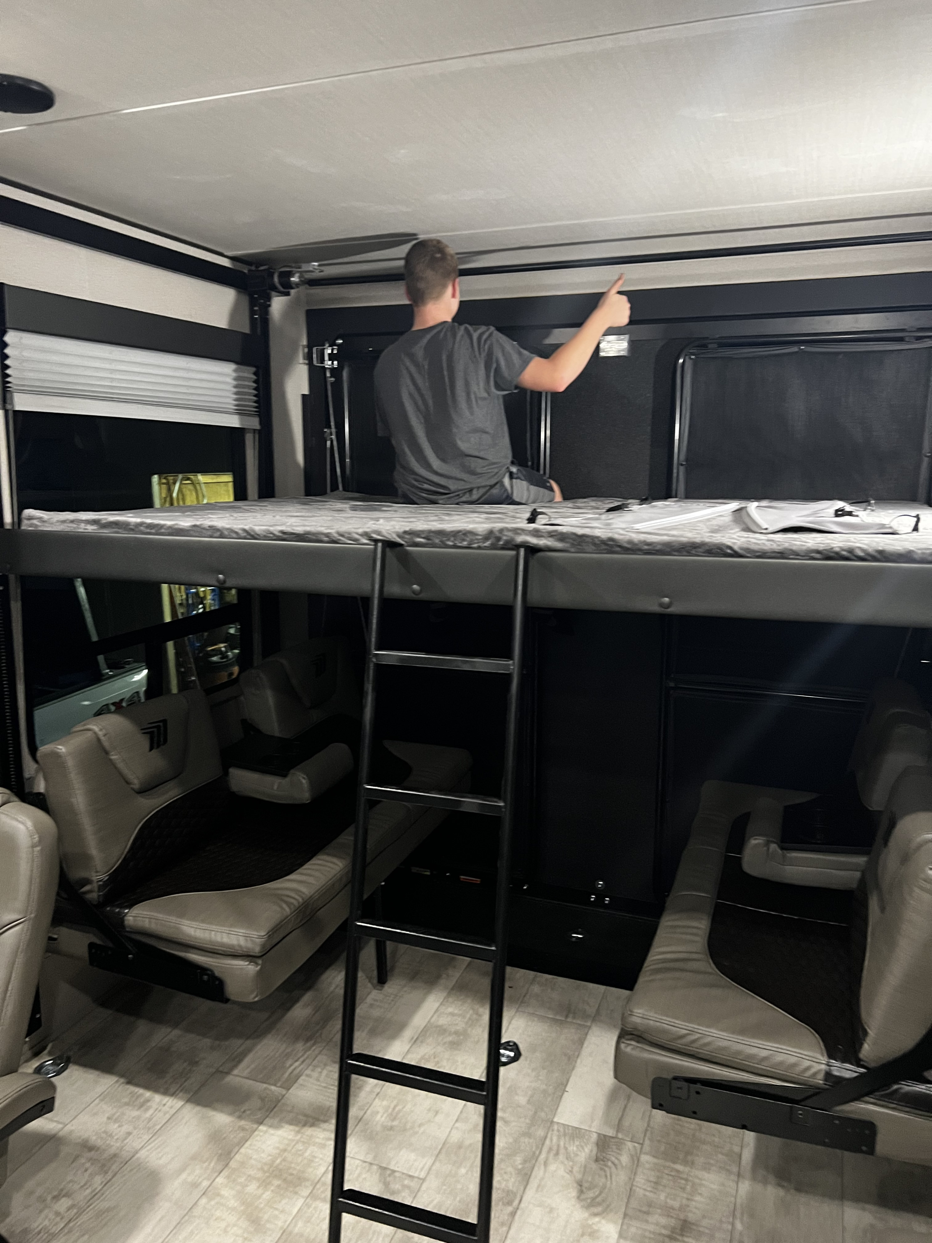 Inside an RV rental, a man sits on a bunk bed, overlooking convertible seating in a toy hauler garage. Ladder provides access.. Grand Design Momentum 2021