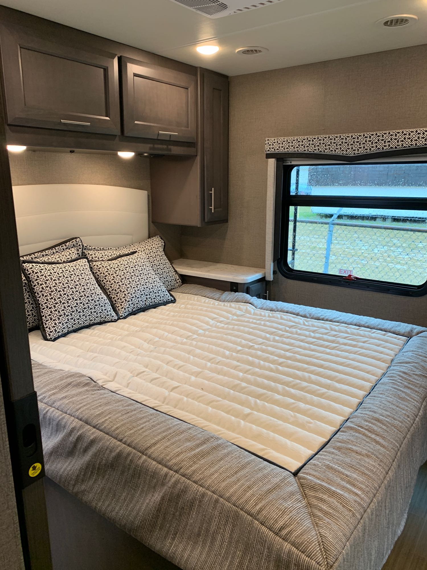 RV rental bedroom with neat bed, white headboard, dark cabinets, and window with exit sign. Compact and organized.. Thor Magnitude 2021