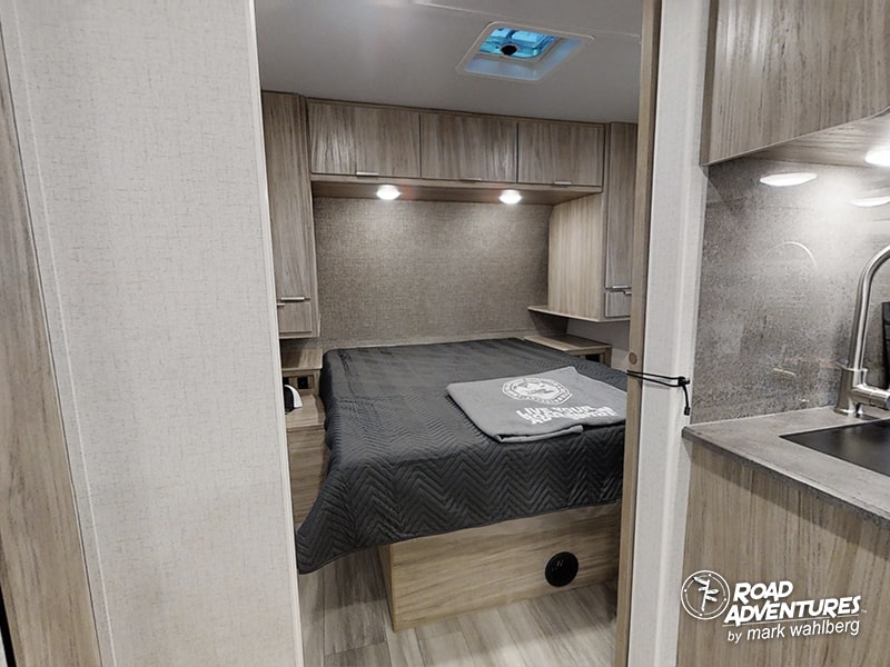 Modern rv rental interior: A compact bedroom with wood cabinets, a gray bed, and a partial kitchen view. Ideal for adventures.. Winnebago Minnie 2301BHS 2022