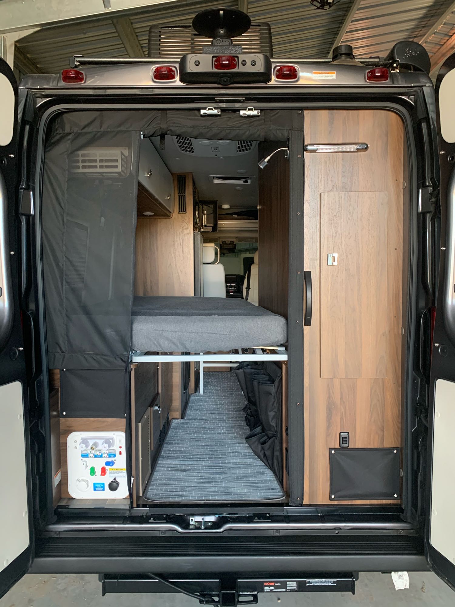 Interior of an RV rental, showing the rear bed, wood cabinetry, and utility panel, viewed from the open back doors.. Winnebago Travato 2022