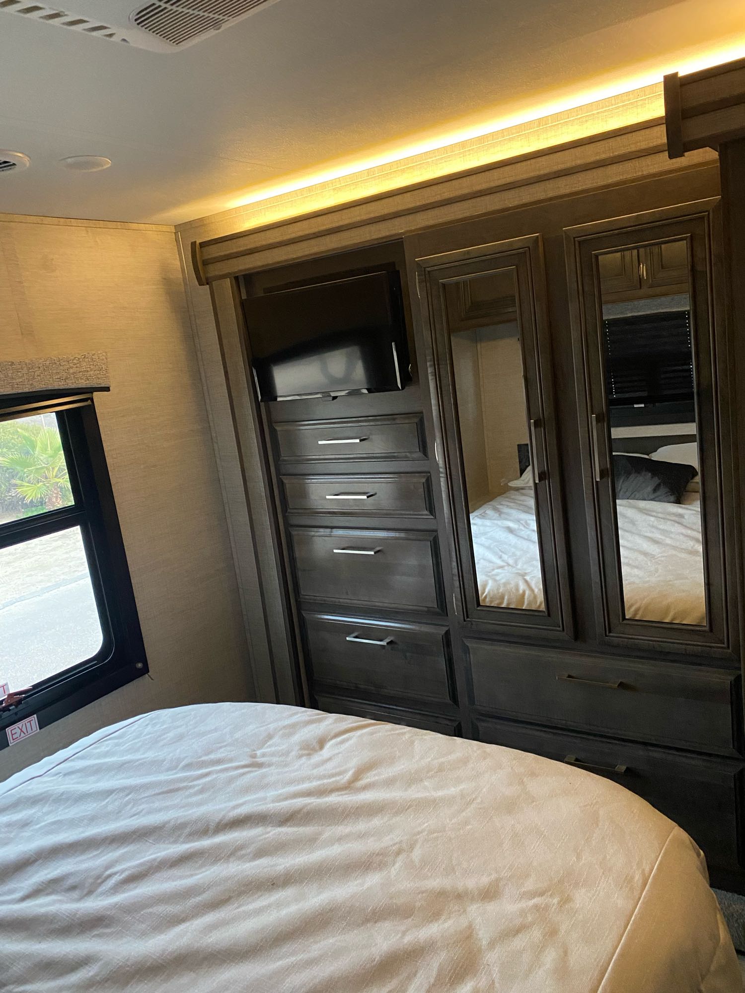 Interior of an RV rental bedroom with a bed, window, and dark wood wardrobe featuring a TV and mirrored doors.. Entegra Coach Odyssey 2021