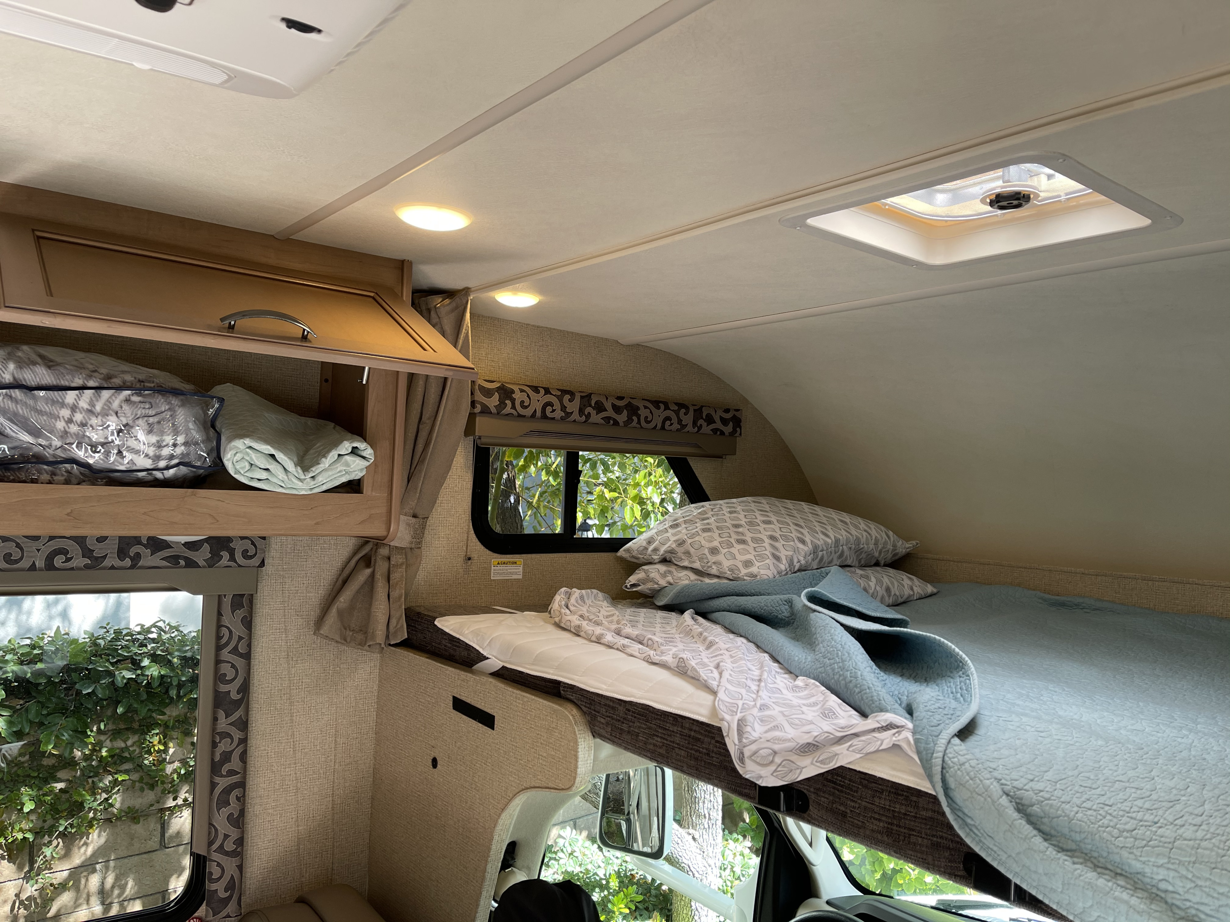 Comfortable cab-over bed area inside an RV rental, showing plush bedding, overhead storage, and scenic windows.. Thor Four Winds 2019