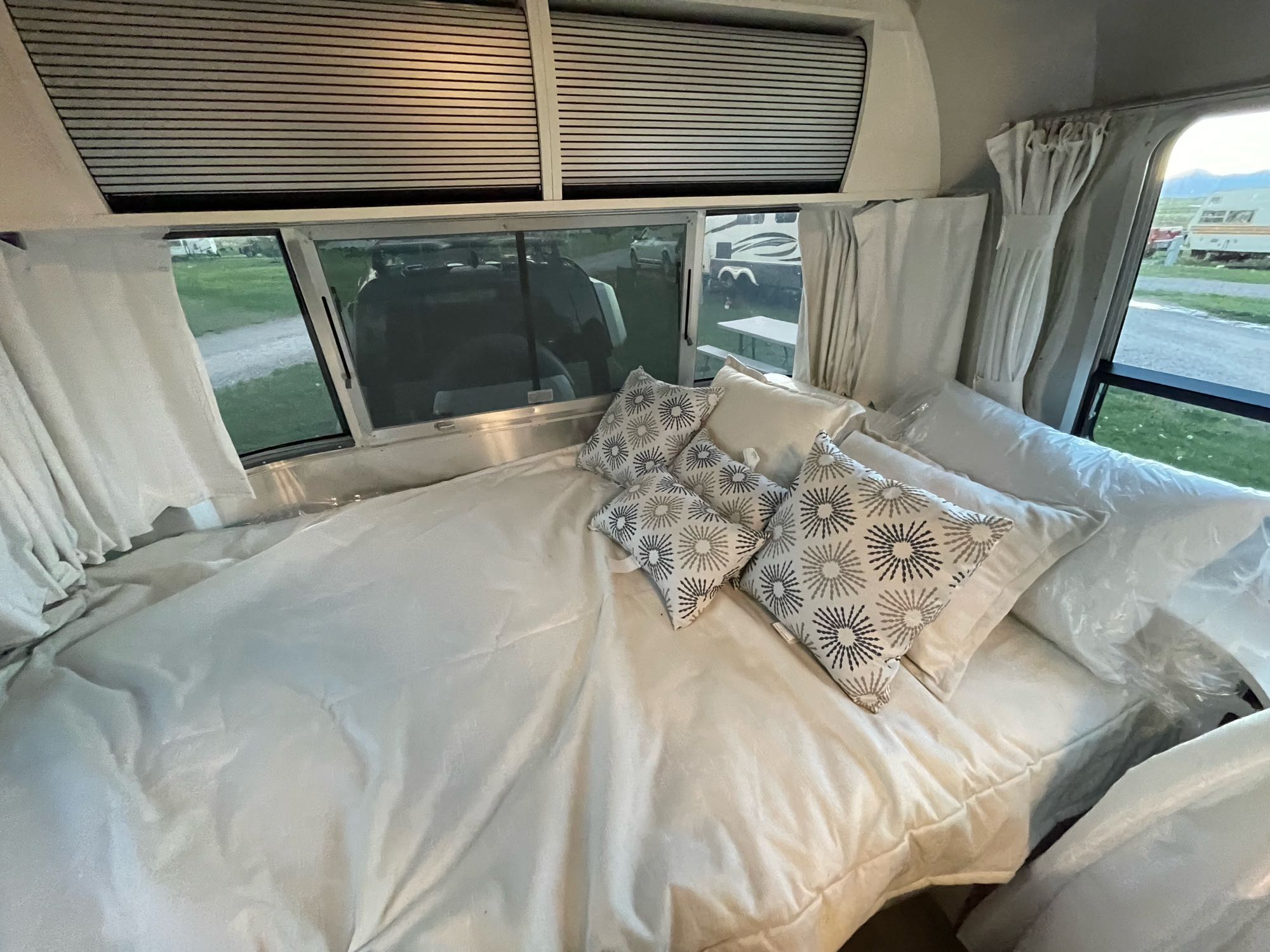 Cozy RV rental bedroom features a comfy bed with white linens and decorative pillows, offering scenic views from large windows.. Airstream Bambi 2021
