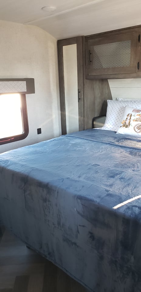 Cozy RV rental bedroom showcasing a plush blue bed, sunlit window, and built-in storage. Ideal for comfortable travel.. Forest River Wildwood 2021