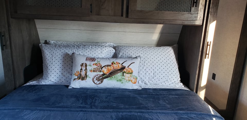 RV rental bedroom view: a cozy bed with polka dot pillows and a decorative fall pillow featuring a dog, pumpkins, and a wheelbarrow.. Forest River Wildwood 2021