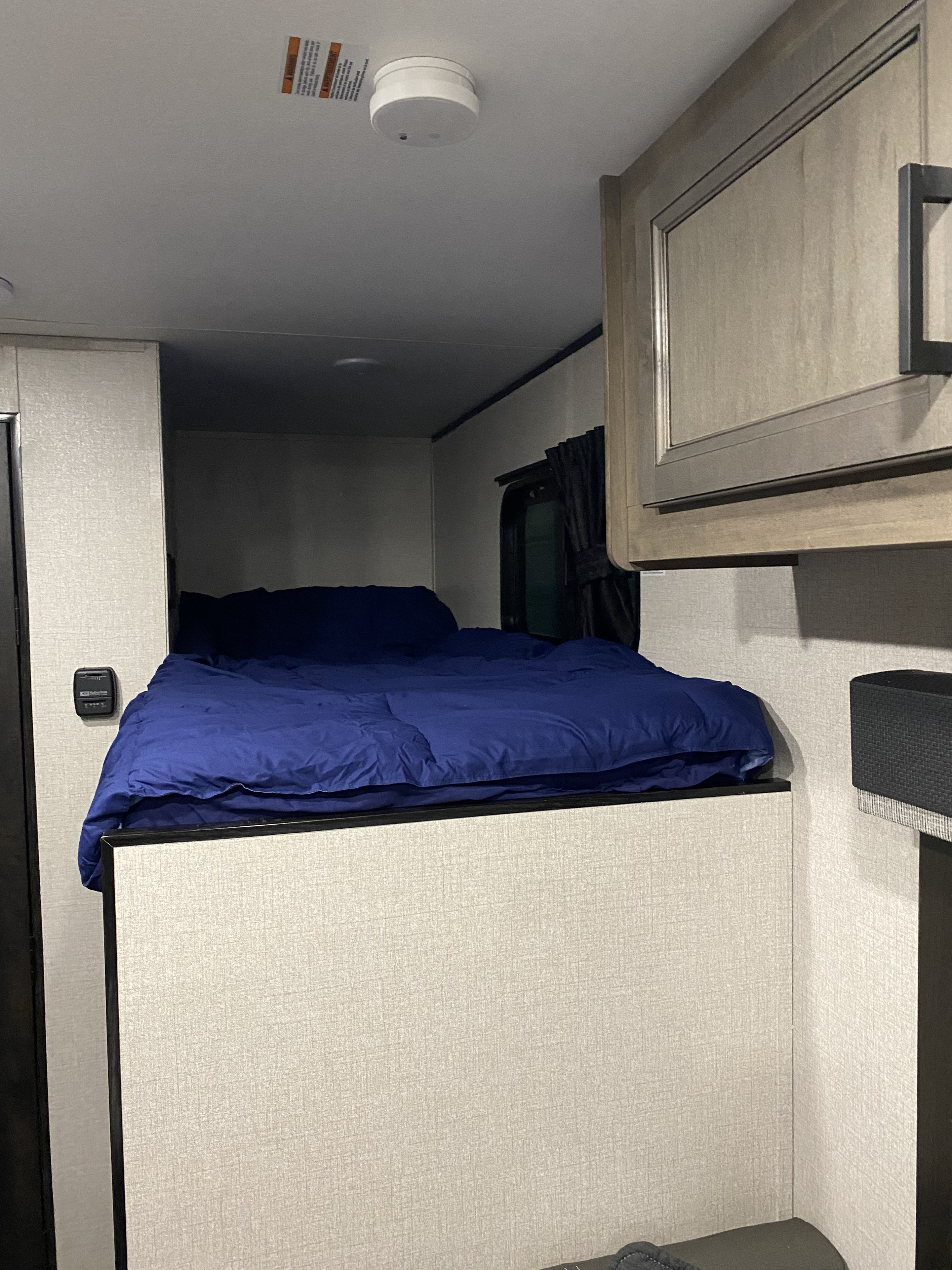 Close-up of an RV rental's cozy bunk bed with blue comforter. Features include an overhead cabinet, thermostat, and ceiling smoke detector.. Highland ridge 19bh 2022