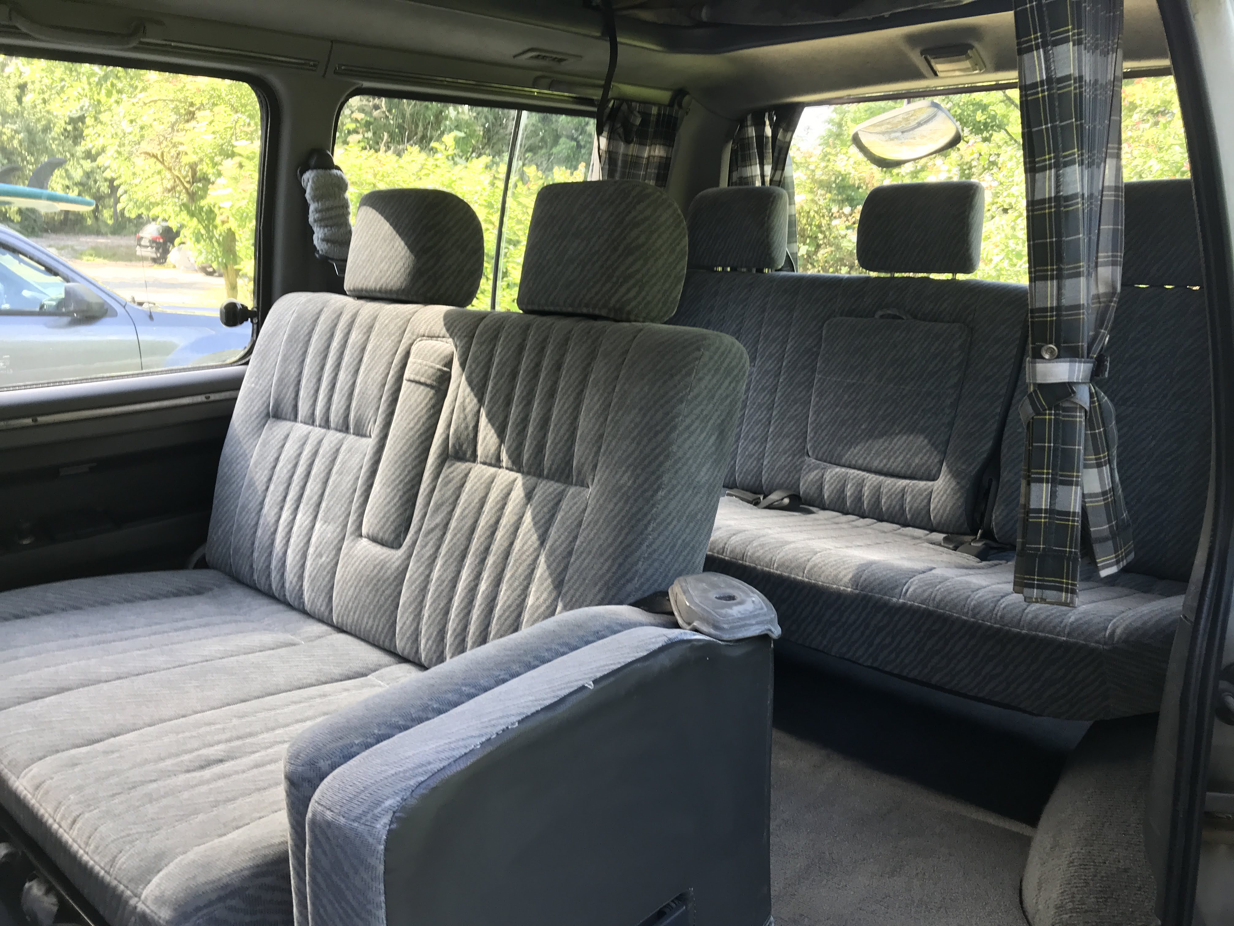 Interior view of an RV rental showing gray striped seats, a convertible bed, and plaid window curtains.. Mitsubishi Delica 1994