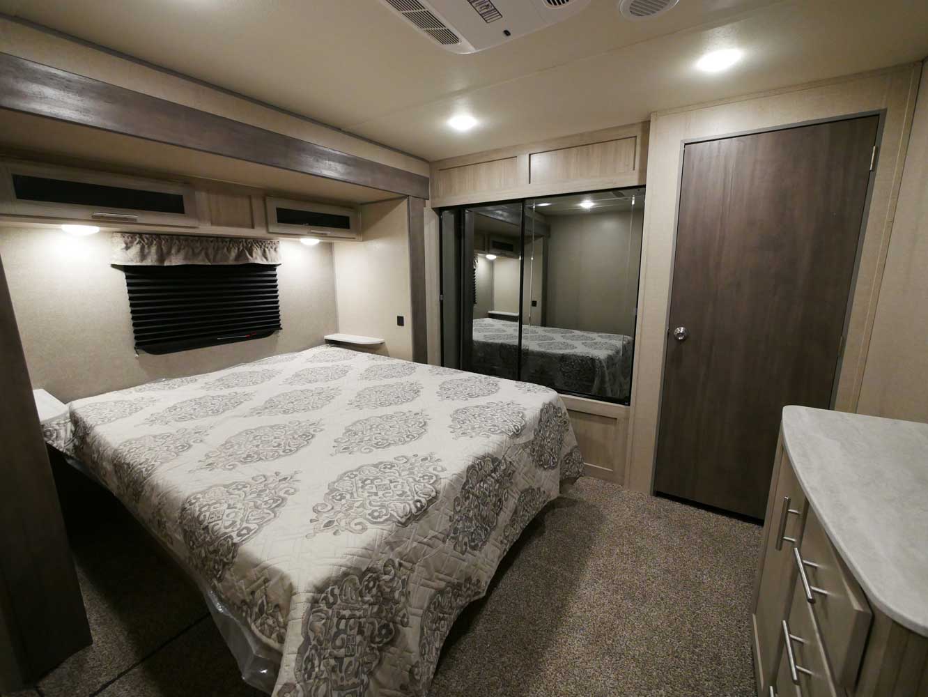 Modern RV rental bedroom featuring a bed with patterned quilt, mirrored wardrobe, dresser, and window.. Forest River Coachmen Catalina Legacy Edition 2022