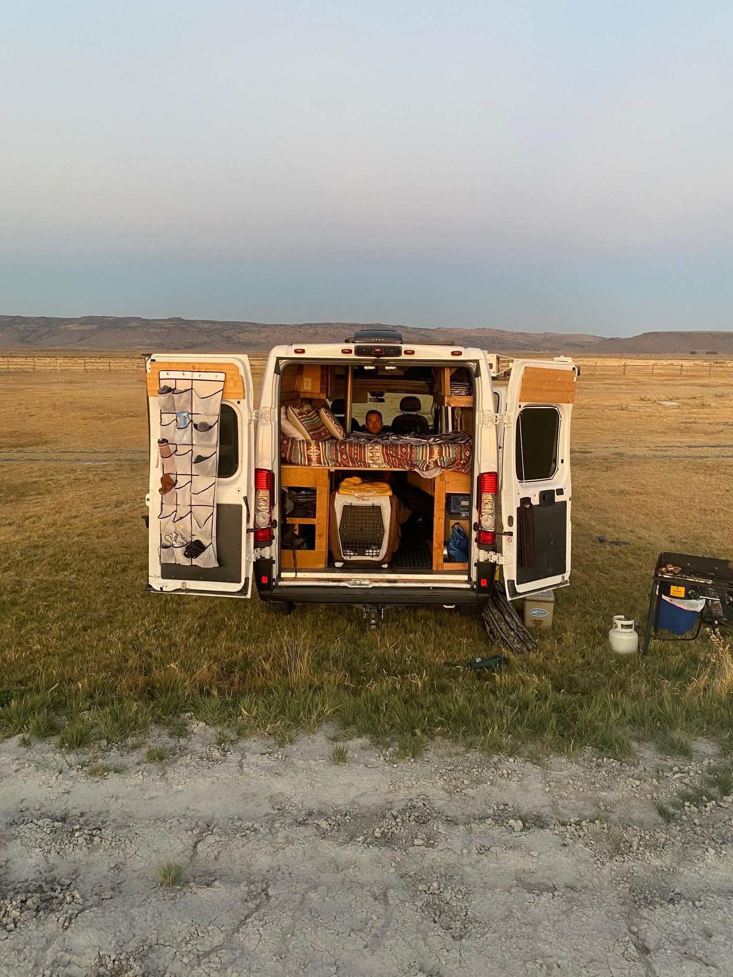 Rear view of an rv rental camper van in a vast field at dusk. Interior shows a person, bed, dog crate, plus outdoor cooking.. Dodge B Van 2019