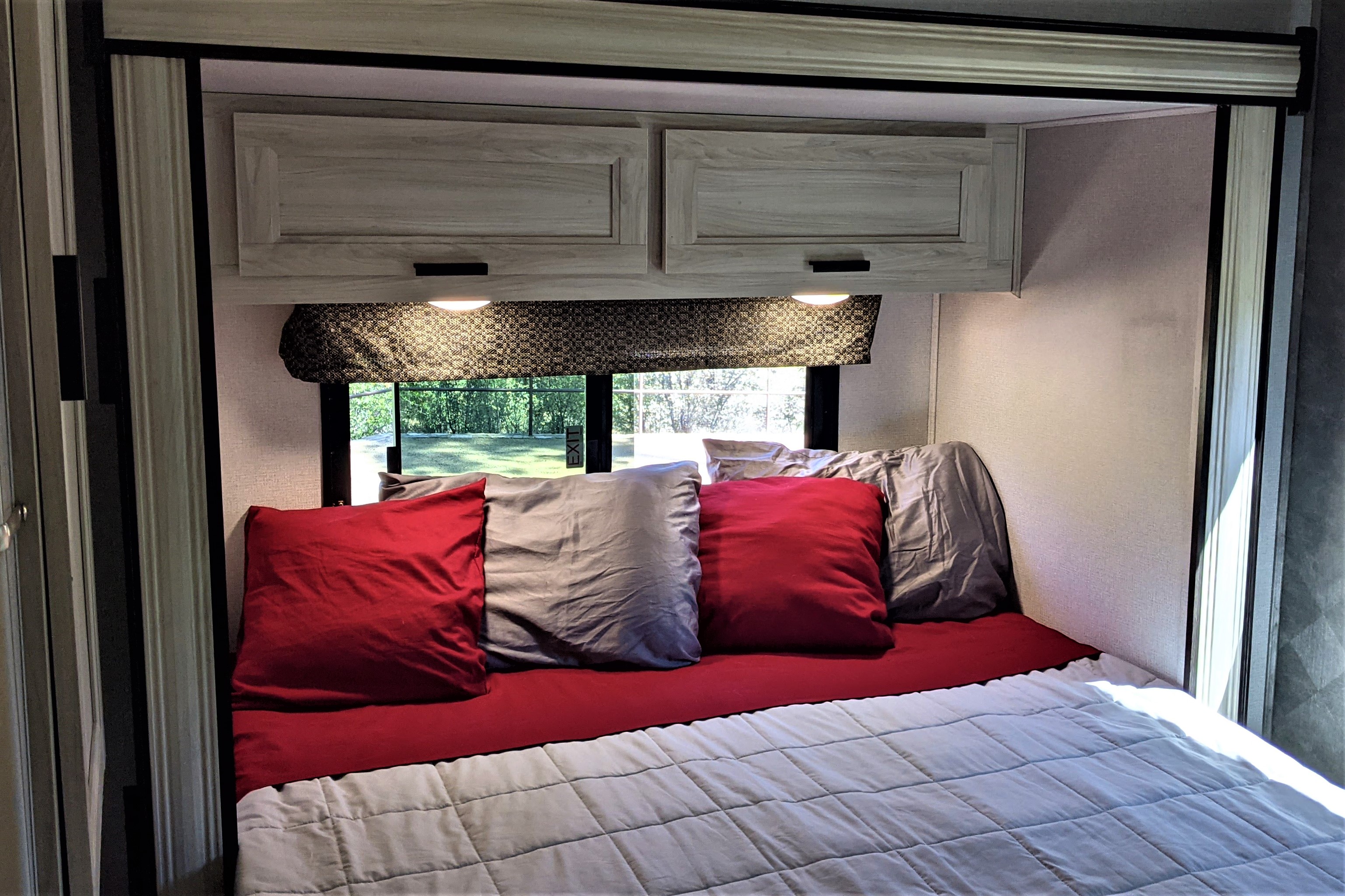 This RV rental bedroom features a comfy bed with red sheets & grey pillows, storage cabinets, and a window with a scenic view.. Forest River Sunseeker LE 2021