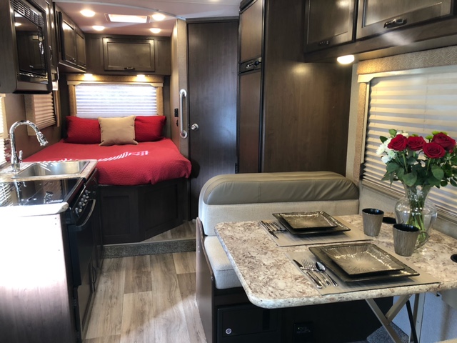 This rv rental interior features a cozy bed, functional kitchenette, and dining booth with flowers on the table.. Thor Freedom Elite 2018