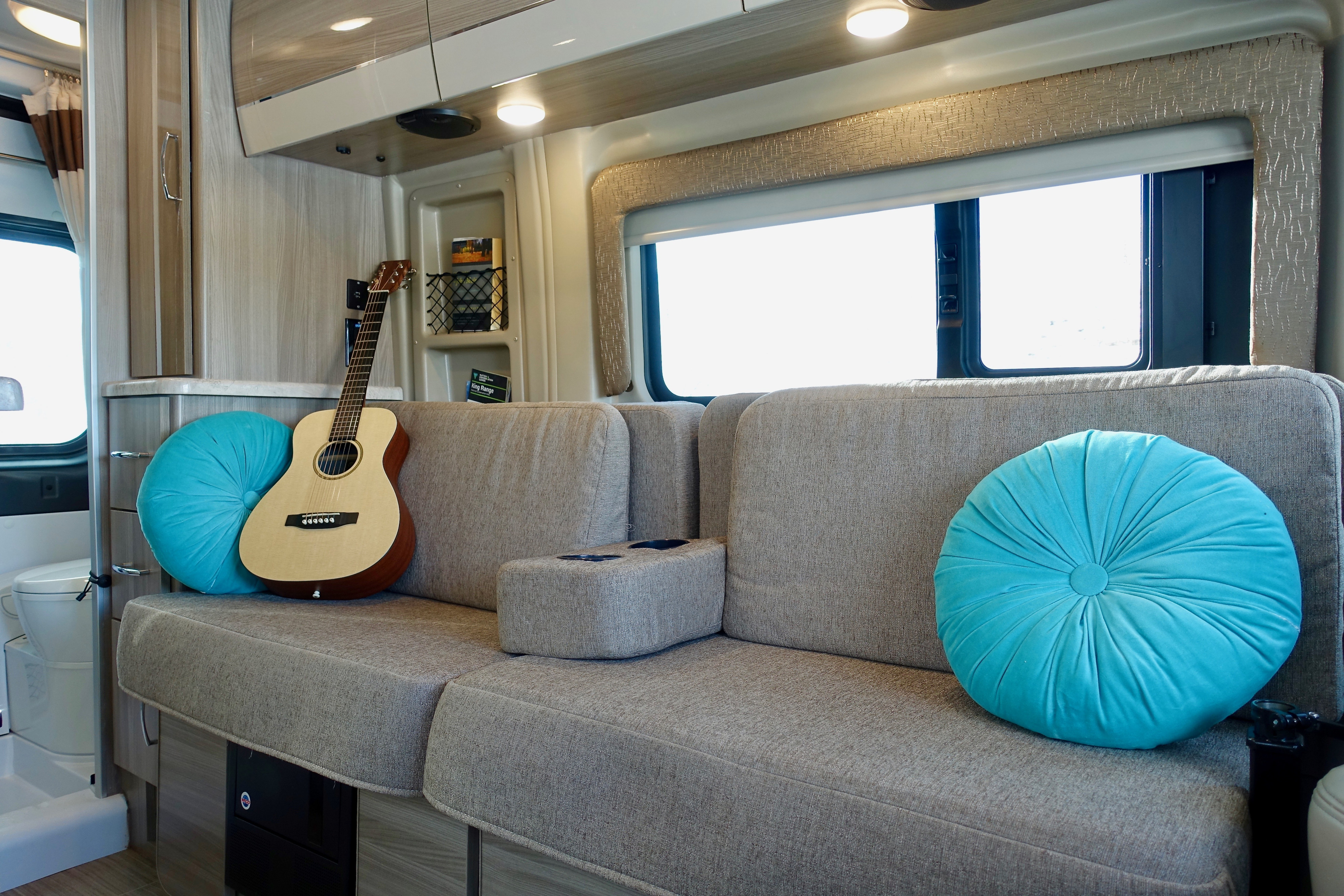 This cozy RV rental living area features a light gray sofa with vibrant turquoise pillows, a guitar, cup holders, and overhead lighting.. Thor Motor Coach Scope 18M 2022