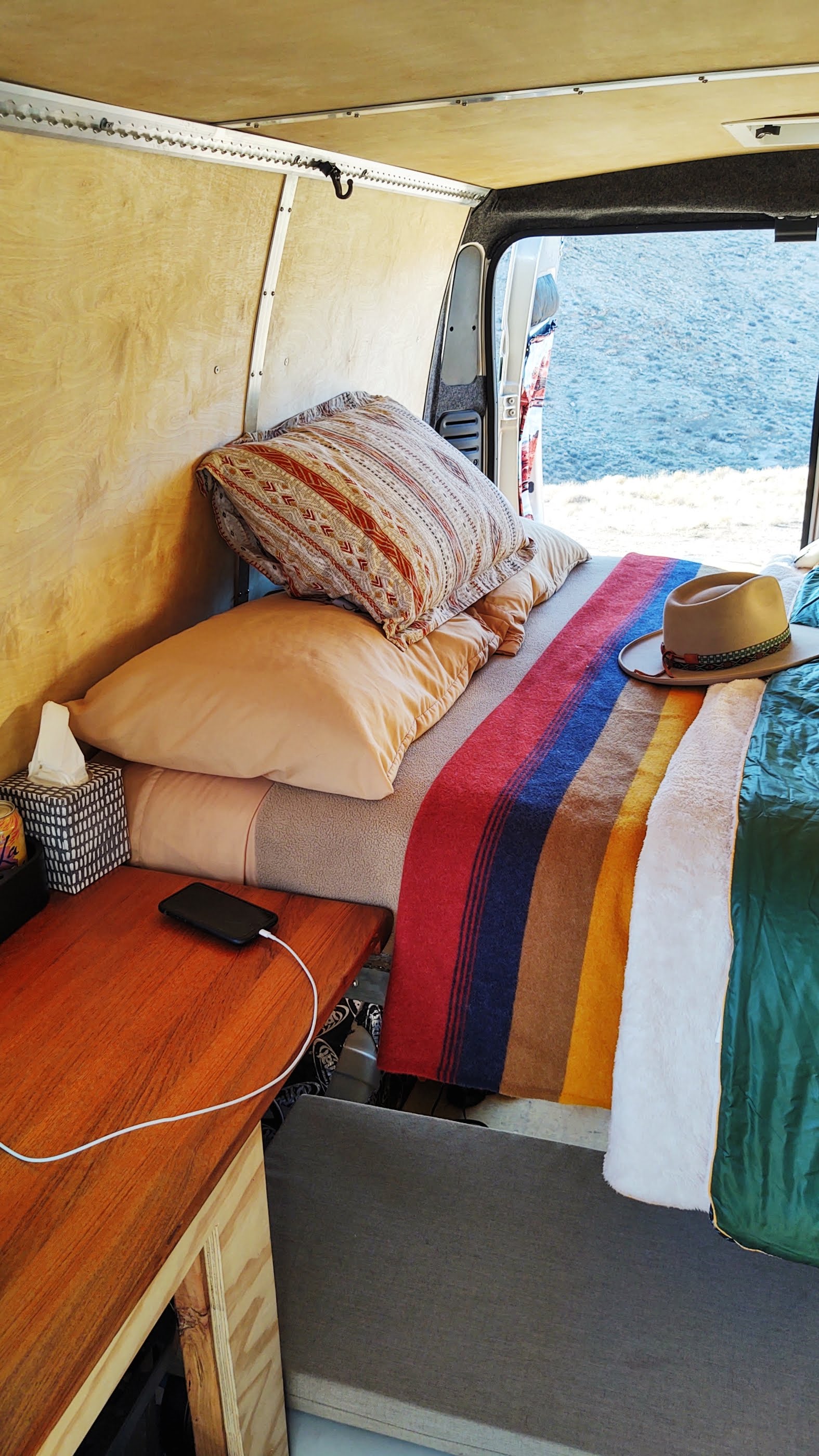 Cozy RV rental bedroom with a colorful blanket, phone charging, and a stunning water view. Perfect for nomadic travel.. Dodge ProMaster 2500 2020