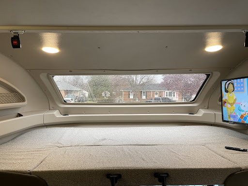 Comfy cab-over bed inside an RV motorhome rental, with a window view of a rainy street and TV entertainment.. Entegra Coach Odyssey 2022